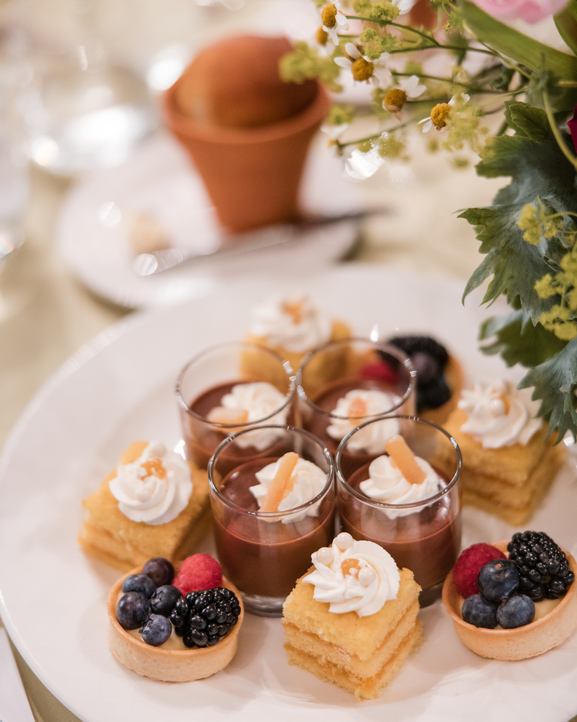 A white plate with assorted desserts—three chocolate mousse cups, three layered cakes with cream topping, and three fruit tartlets—served beside a floral arrangement at the Longwood Gardens welcome party.