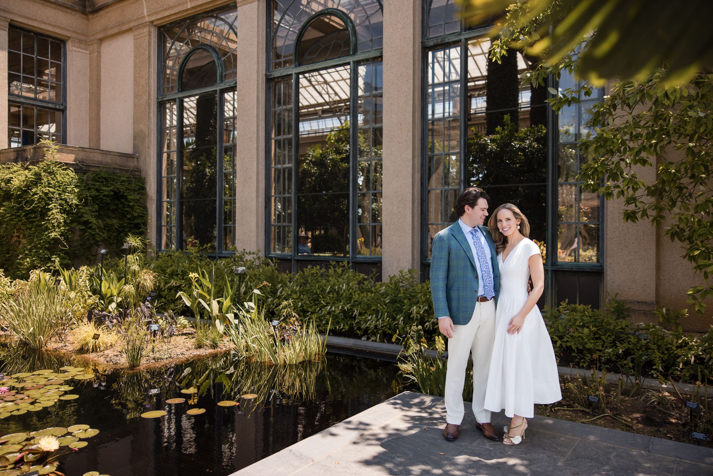 A couple stands near a pond in front of a building with large glass windows, enjoying the ambiance of the Longwood Gardens welcome party.