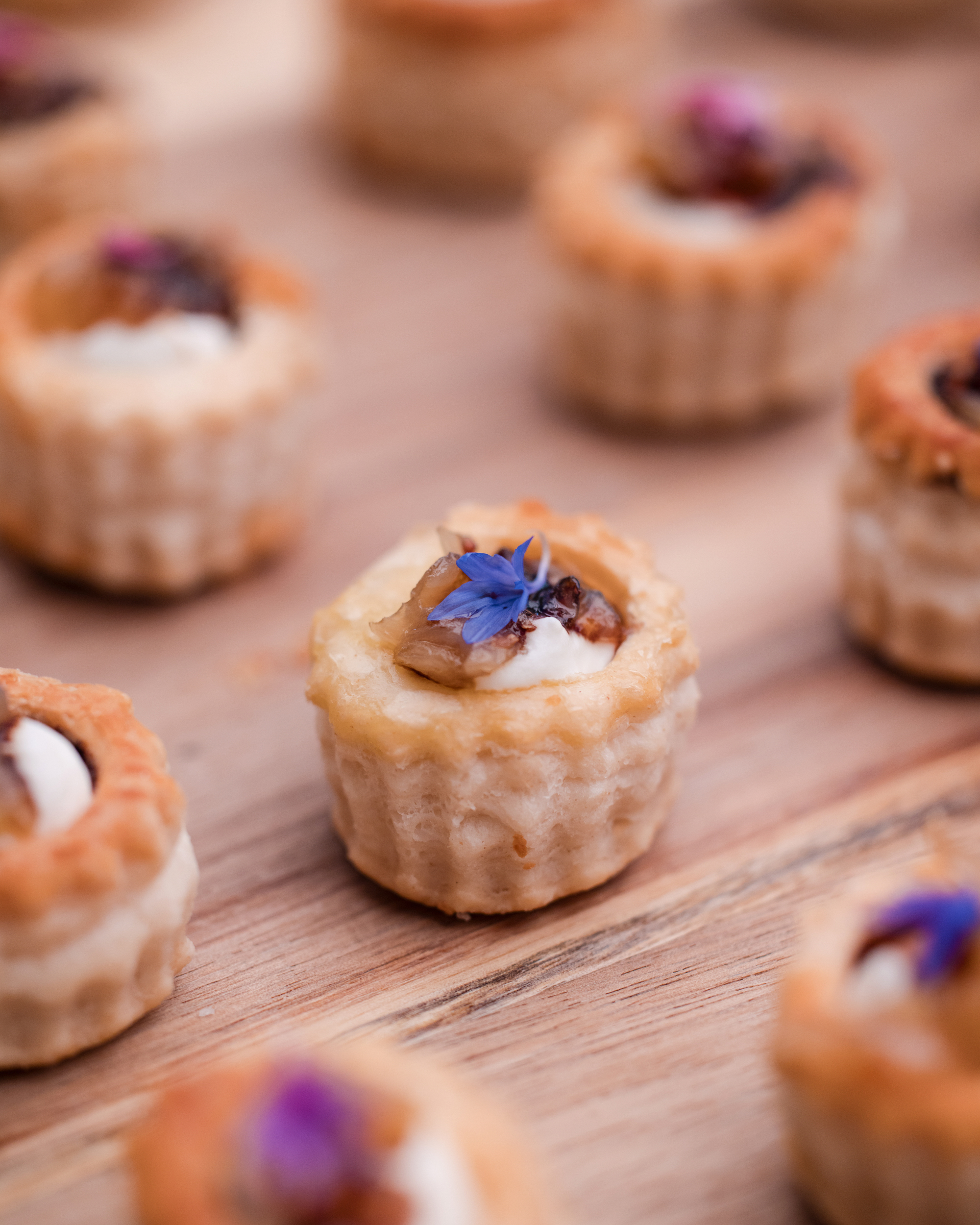 Mini pastry tartlets with creamy filling, topped with walnuts and blue edible flowers, arranged on a wooden surface—perfect bites for a Longwood Gardens welcome party.