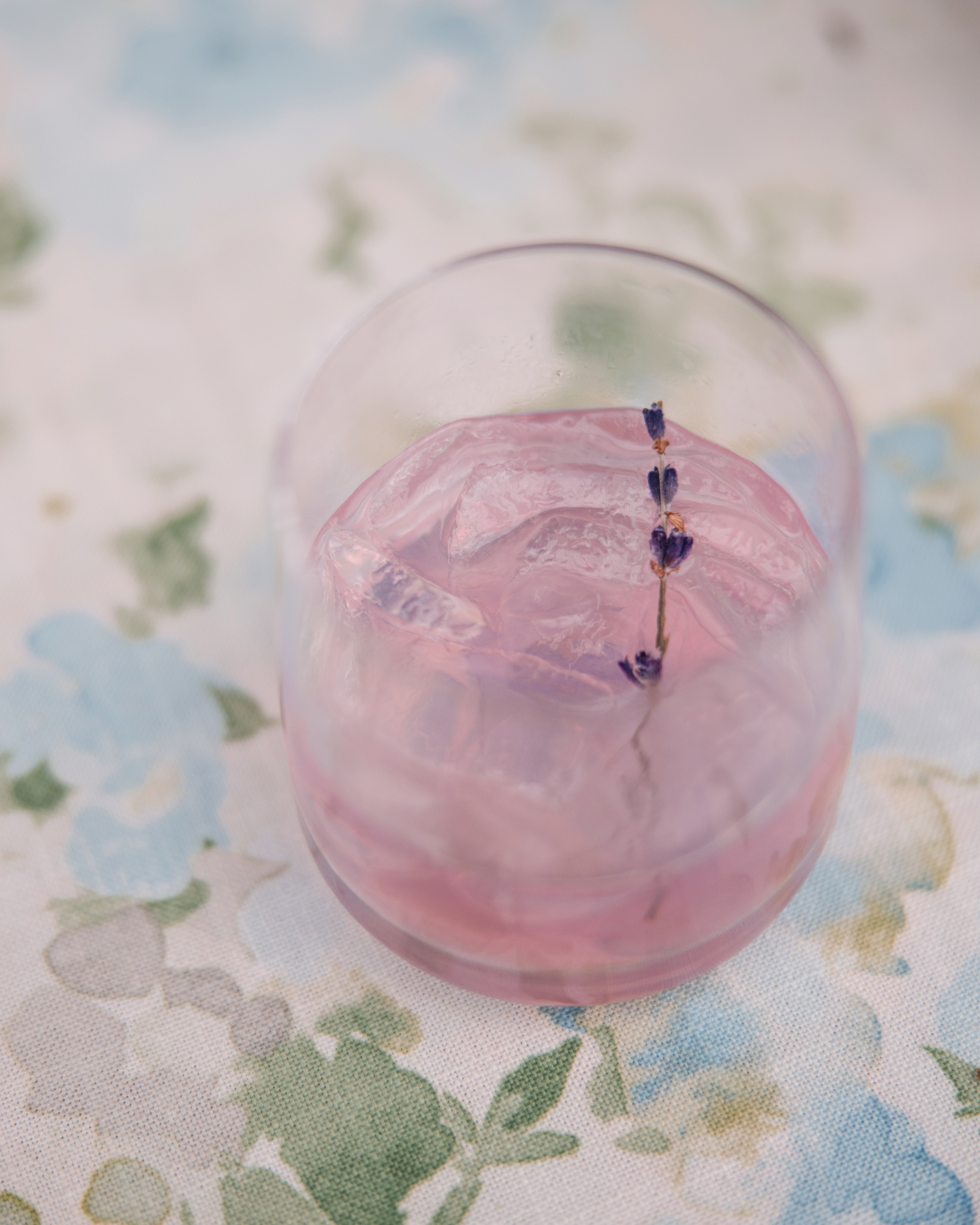 A glass of pink beverage with ice, garnished with a small sprig of lavender, sits on a floral-patterned tablecloth—perfect for a Longwood Gardens welcome party.
