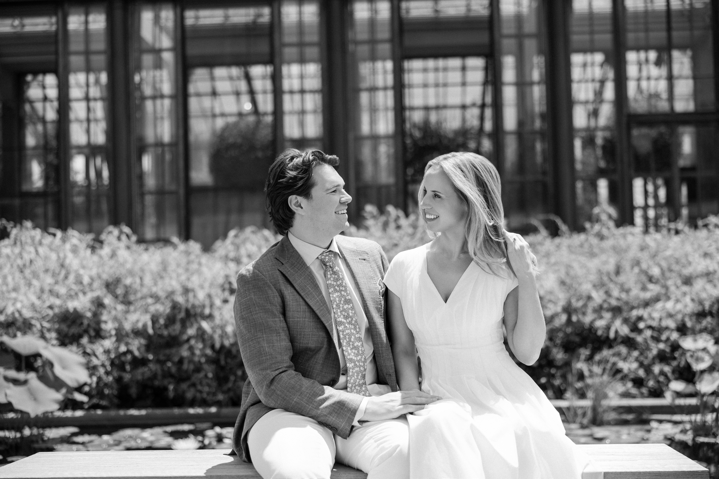 A man in a suit and a woman in a white dress sit together on a bench outside at Longwood Gardens, greenery and a building behind them—capturing a quiet moment before the welcome party begins.
