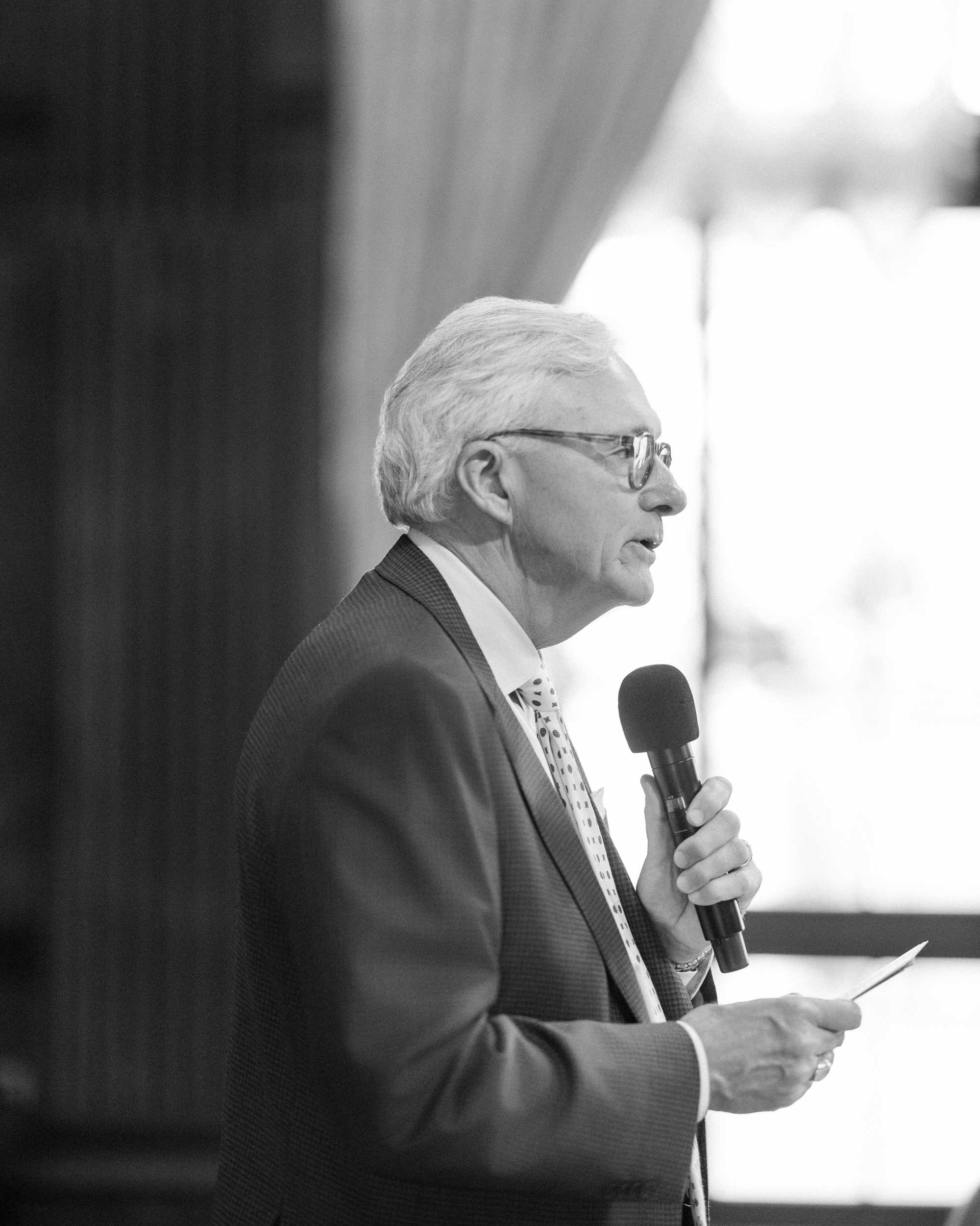 At a Longwood Gardens welcome party, a man in a suit and tie speaks into a microphone while holding a piece of paper, standing indoors near a window with curtains.