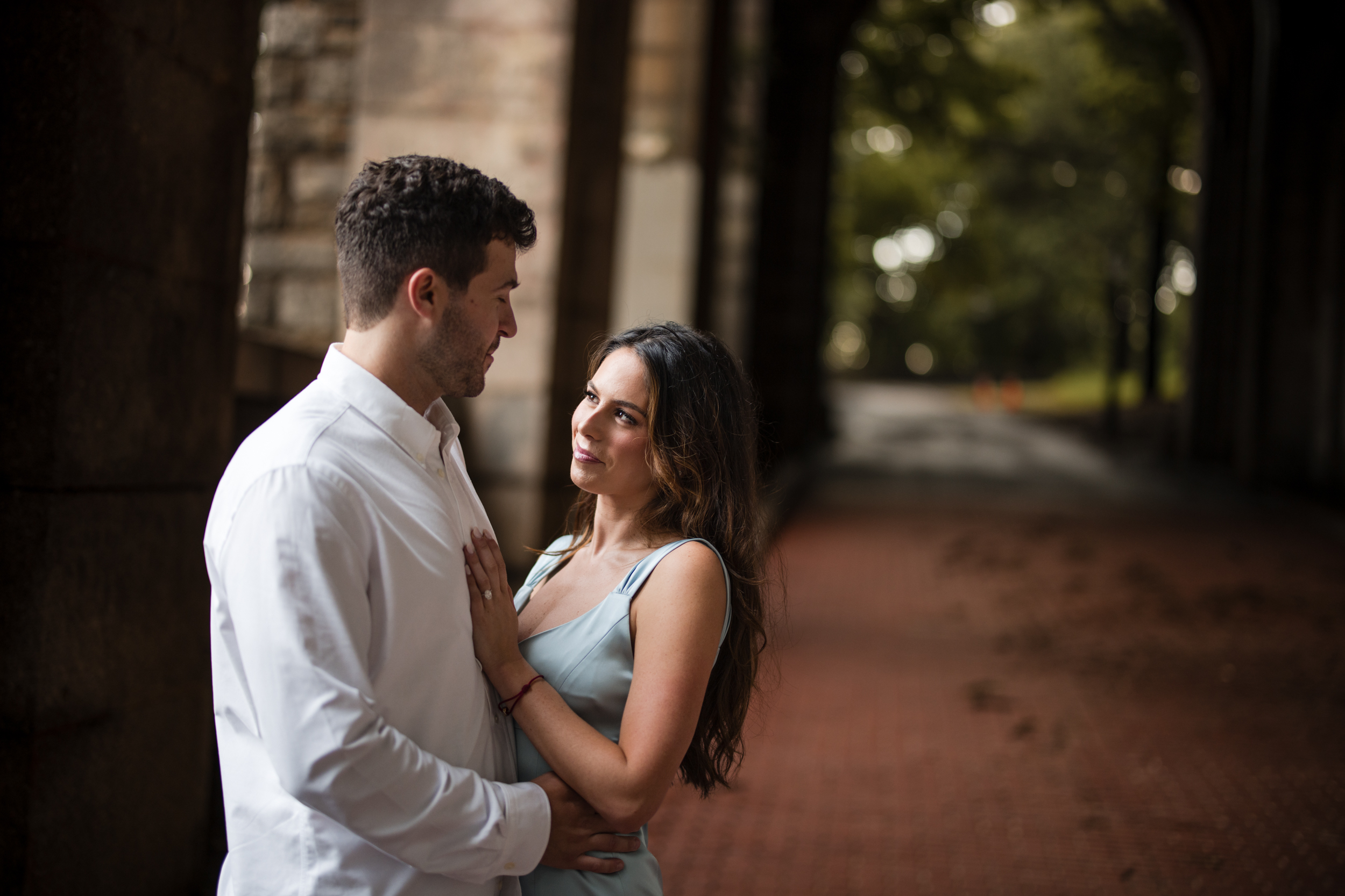 A man and woman stand close together under an archway, looking at each other. The background, typical of Fort Tryon Park engagement photos, shows a tree-lined path and blurred greenery.