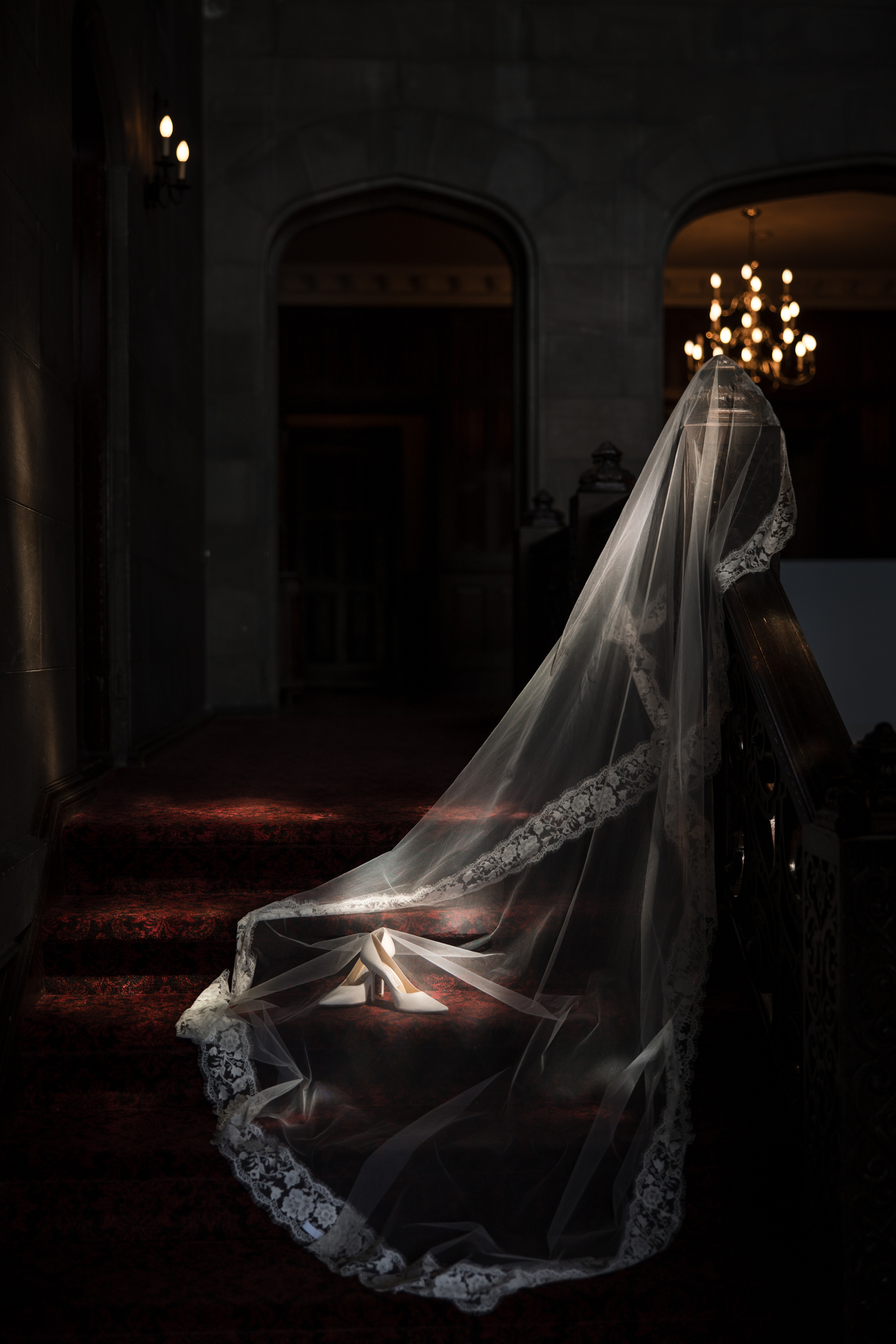 A bride wearing a long lace veil stands on a dimly lit staircase under a chandelier at Hempstead House at Sands Point Preserve, with light highlighting her veil and shoes.