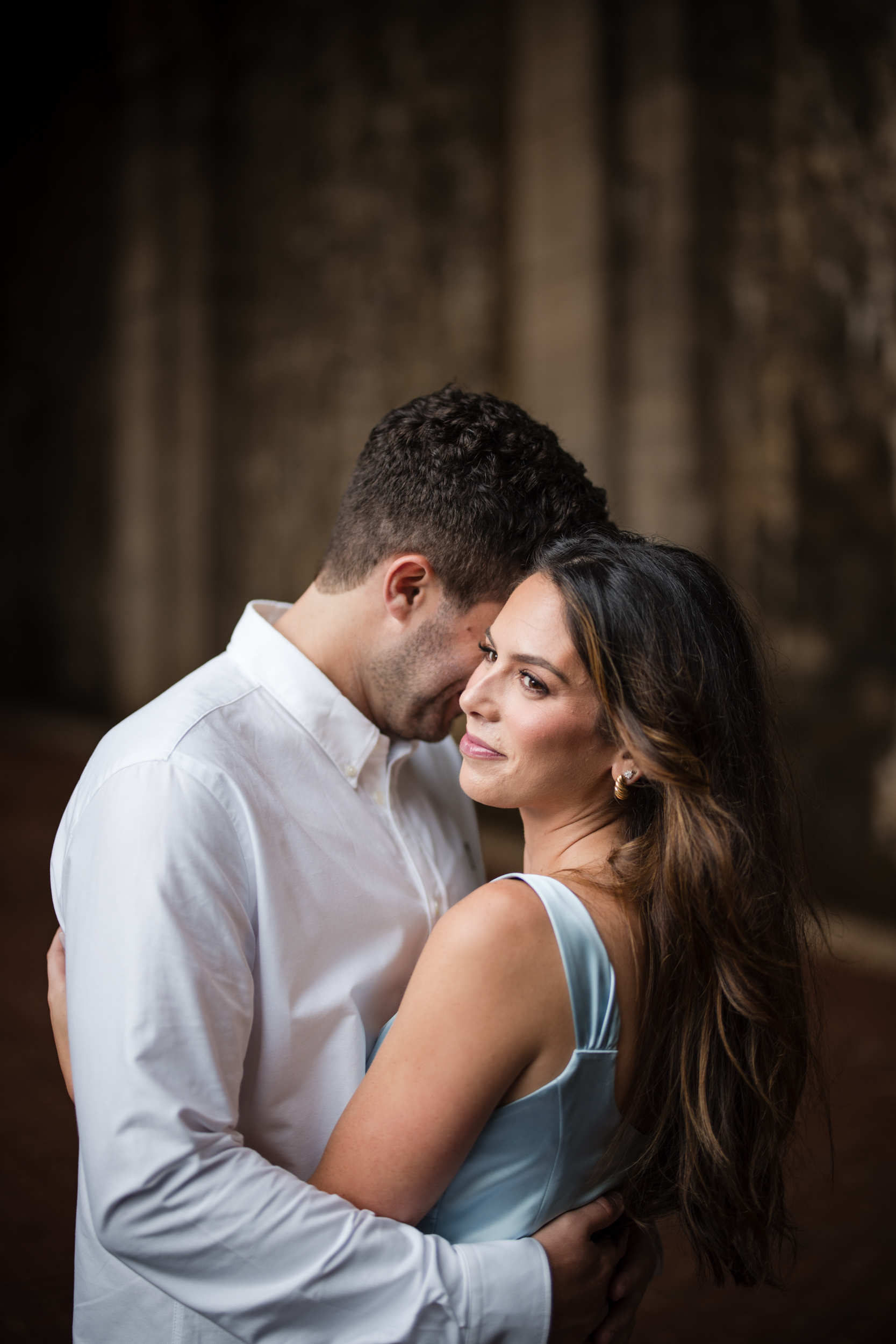 A woman in a light blue dress smiles while being embraced by a man in a white shirt against a blurred, dark stone background, capturing the romantic essence of Fort Tryon Park engagement photos.