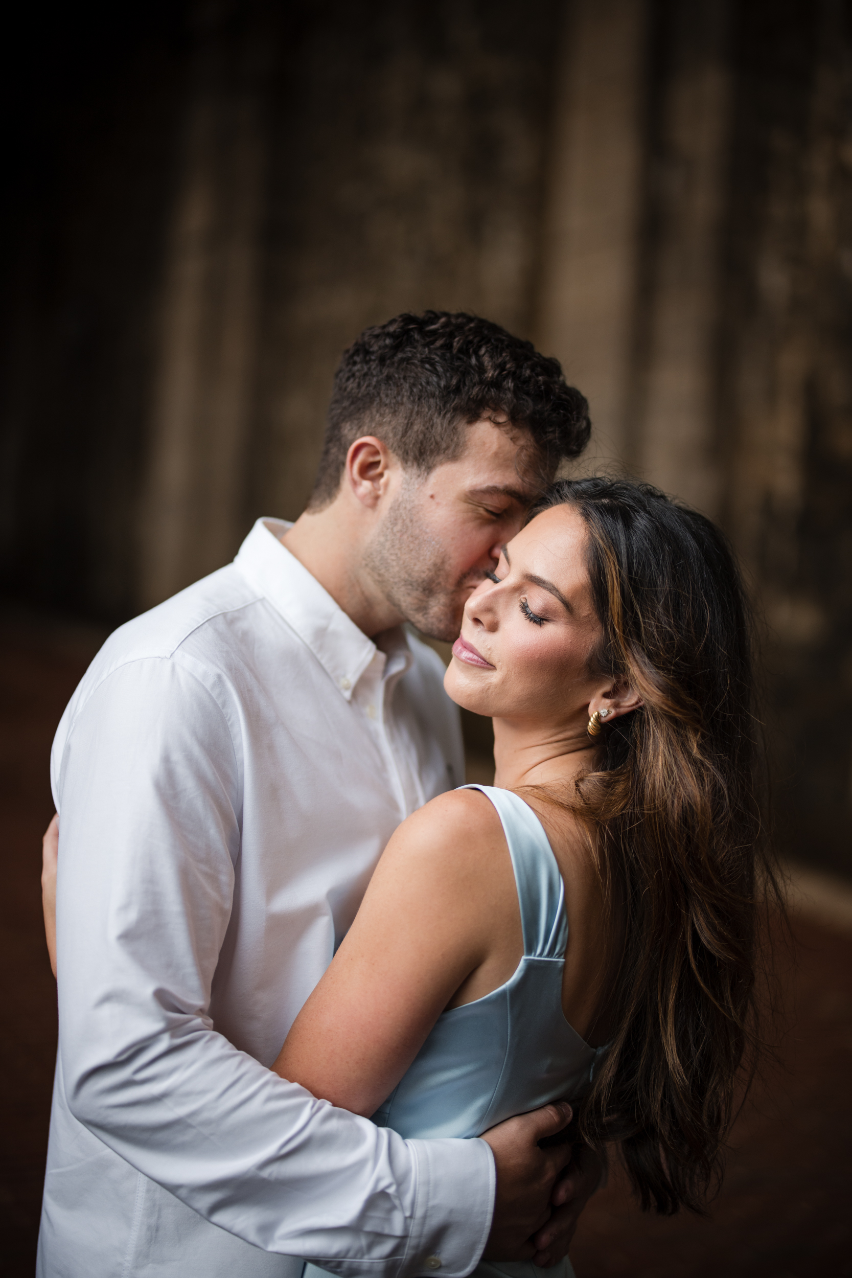 A man in a white shirt embraces a woman in a light blue dress, gently kissing her on the forehead. They stand close together with a blurred, dark background, capturing an intimate moment reminiscent of Fort Tryon Park engagement photos.