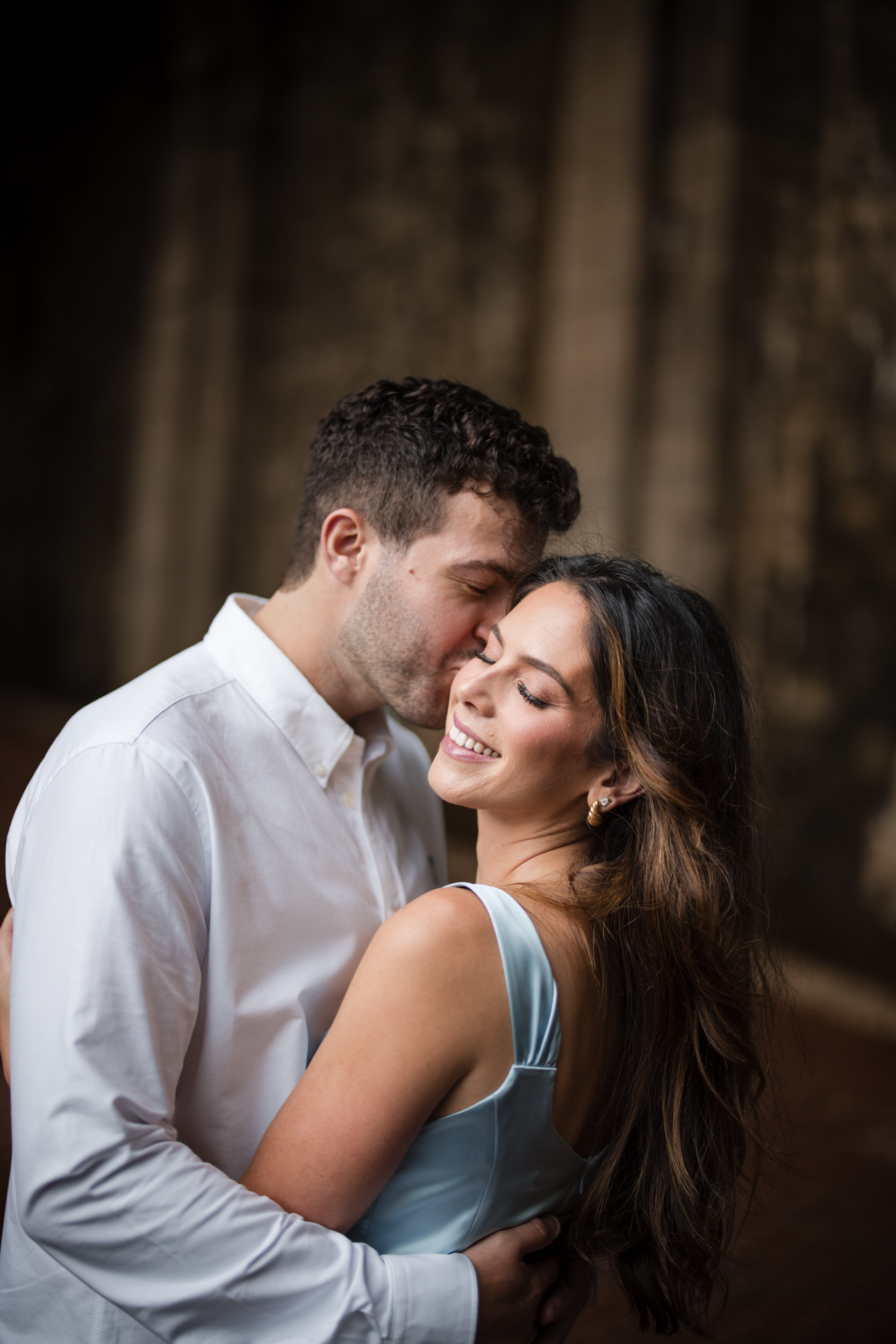 A man in a white shirt kisses a smiling woman in a light blue dress on the forehead as they embrace in front of a blurred brown background, capturing the tender moment during their Fort Tryon Park engagement photos.