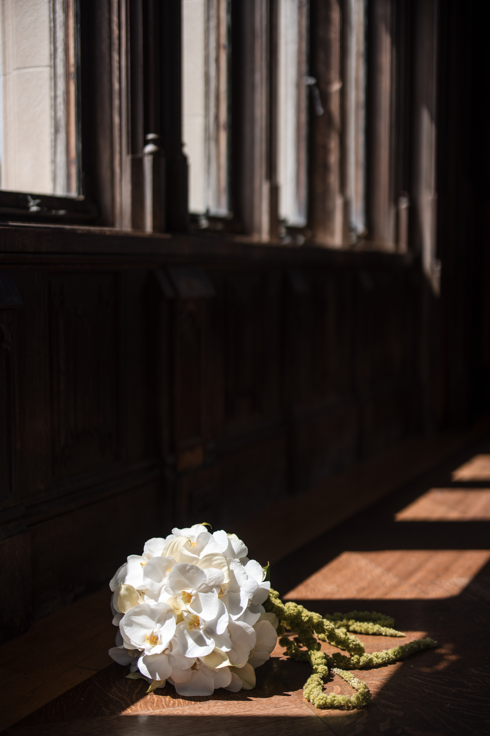 A bouquet of white flowers with green stems lies on a sunlit wooden floor, next to a dark wood-paneled wall and tall windows—capturing the timeless elegance of a Hempstead House at Sands Point Preserve wedding.