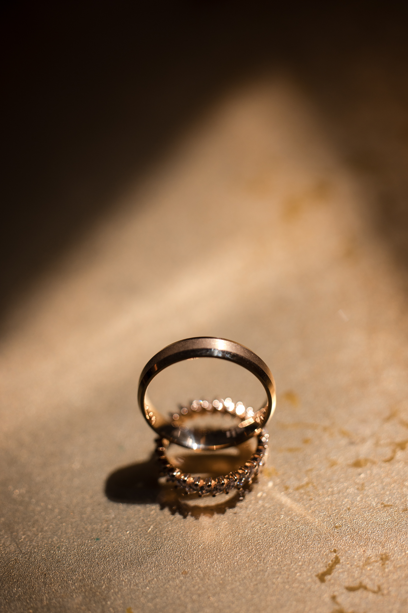 Two gold rings, one plain and one with small stones, are standing upright on a textured surface in warm, soft lighting—capturing the timeless romance of a Hempstead House at Sands Point Preserve wedding.