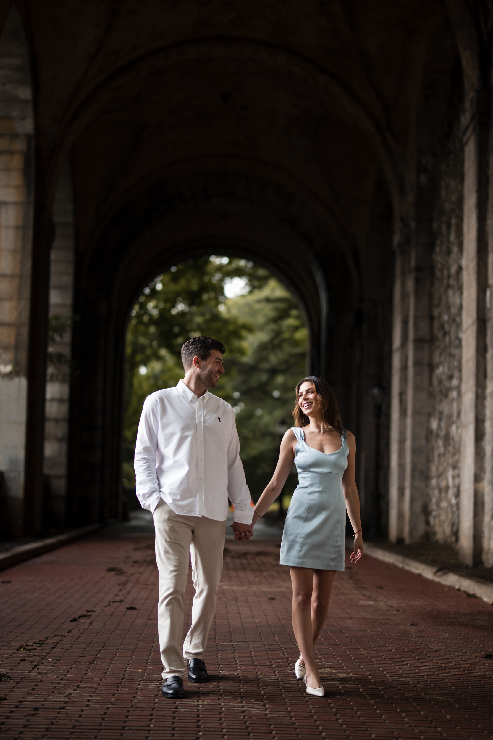 A man and woman walk hand in hand on a brick path under a large stone archway, both smiling and looking at each other, capturing a romantic moment during their Fort Tryon Park engagement photos.