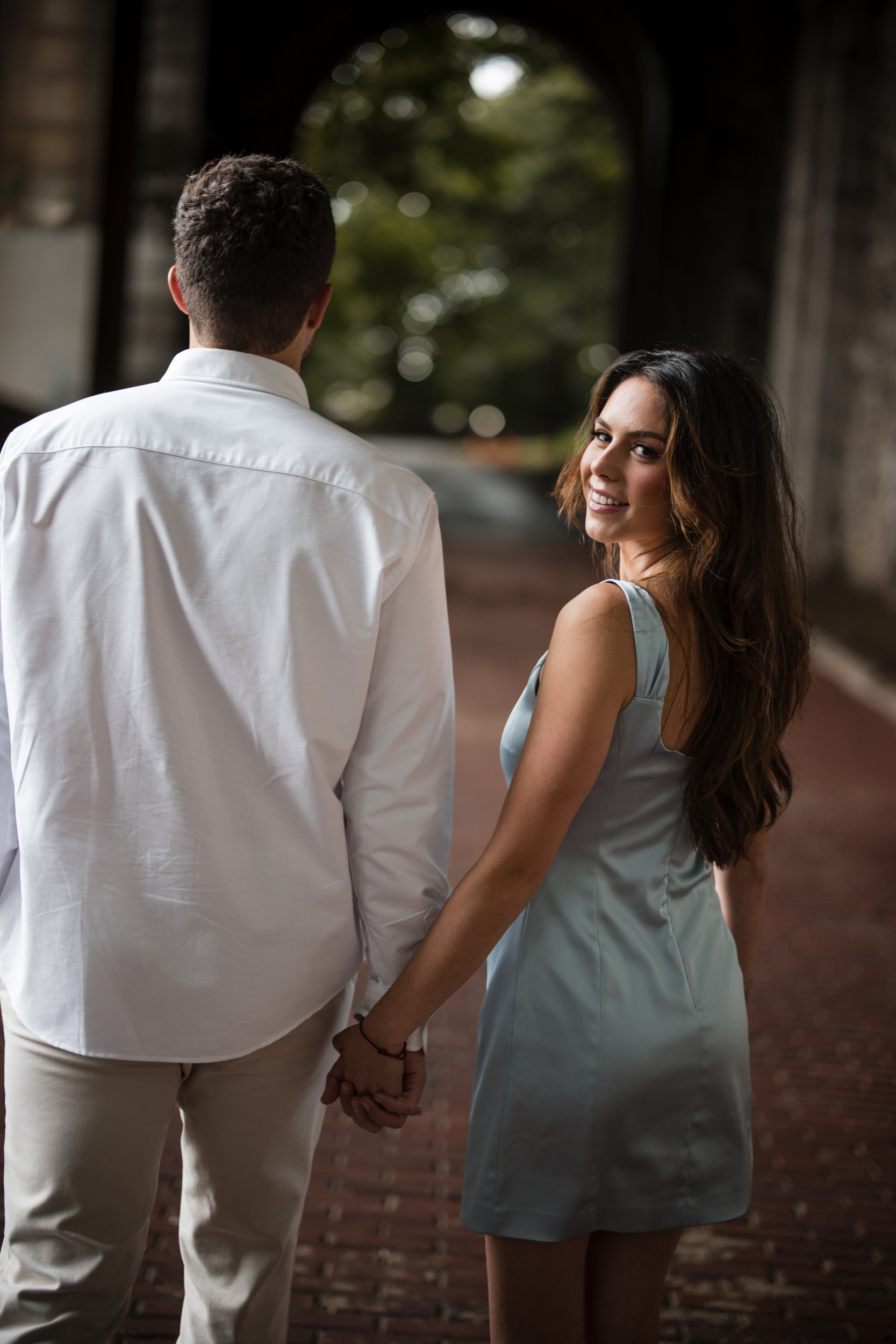 Woman in a light blue dress smiles at the camera while holding hands with a man in a white shirt as they walk down a brick path under an archway, capturing the perfect Fort Tryon Park engagement photos.