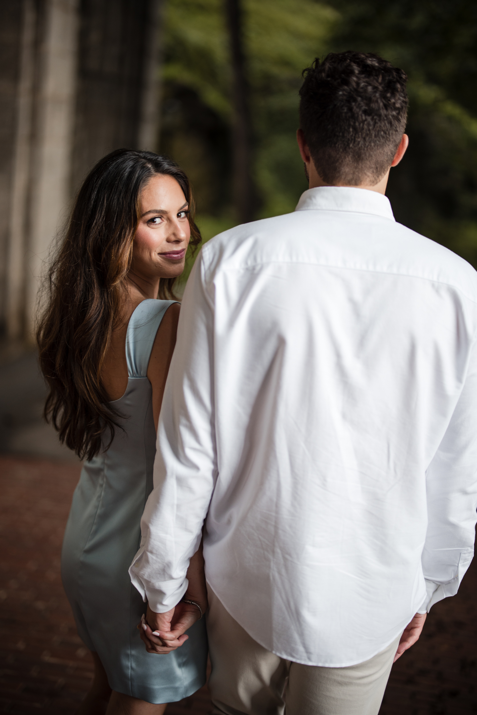 A woman in a light blue dress holds hands with a man in a white shirt, looking over her shoulder and smiling. They are outdoors near a stone structure, capturing the romantic charm of Fort Tryon Park engagement photos.