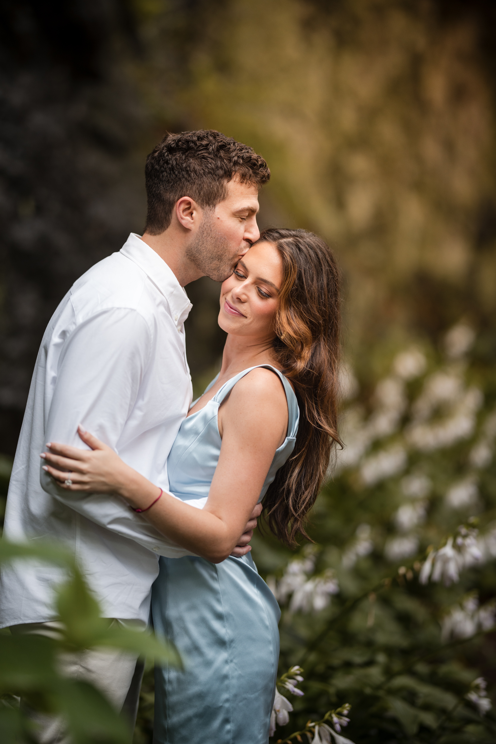 A man in a white shirt kisses a woman in a light blue dress on the forehead as they embrace in a beautiful garden setting during their Fort Tryon Park engagement photos.