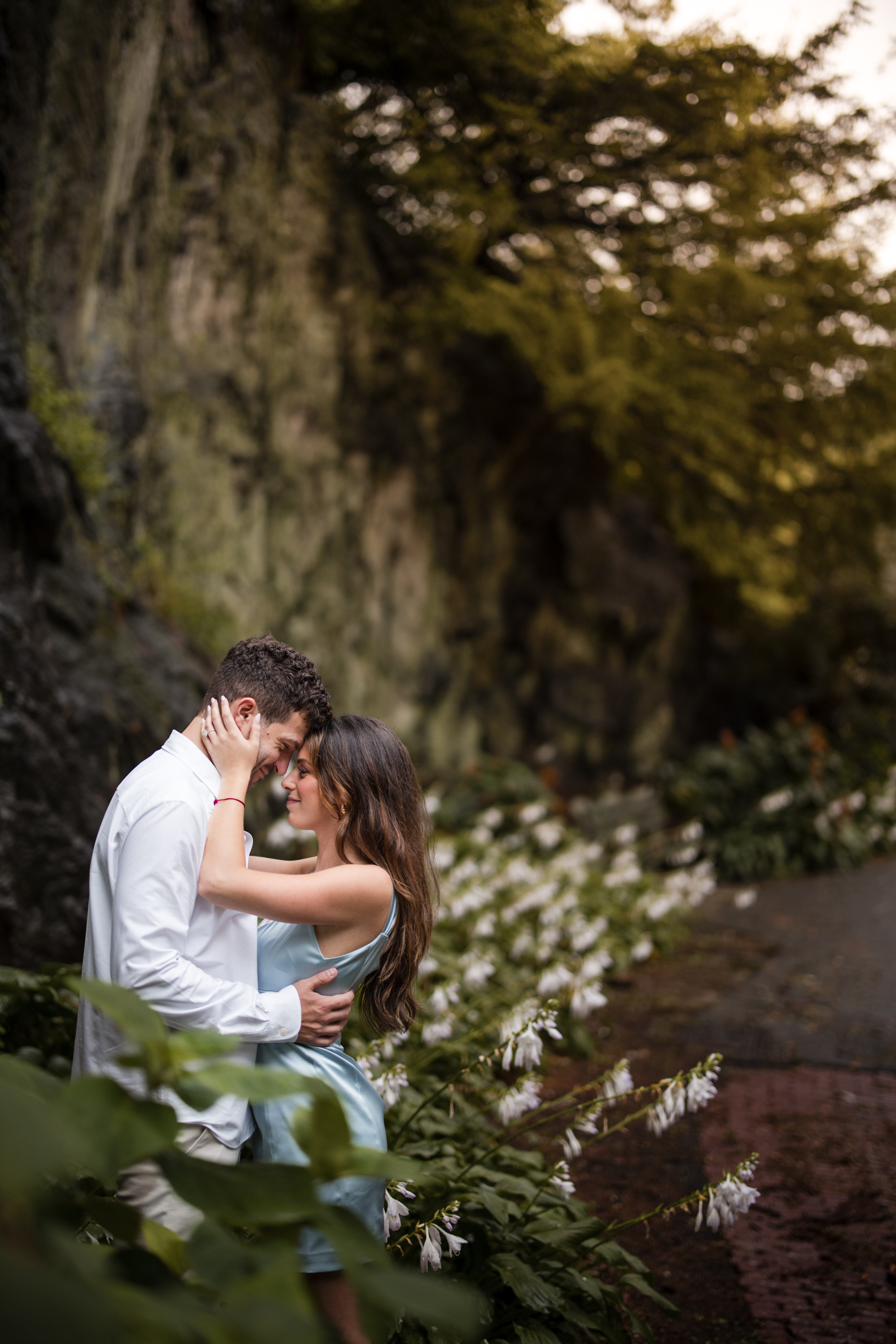 A couple stands close together, embracing on a path lined with green plants and white flowers near a rocky wall, capturing the romance of their Fort Tryon Park engagement photos.