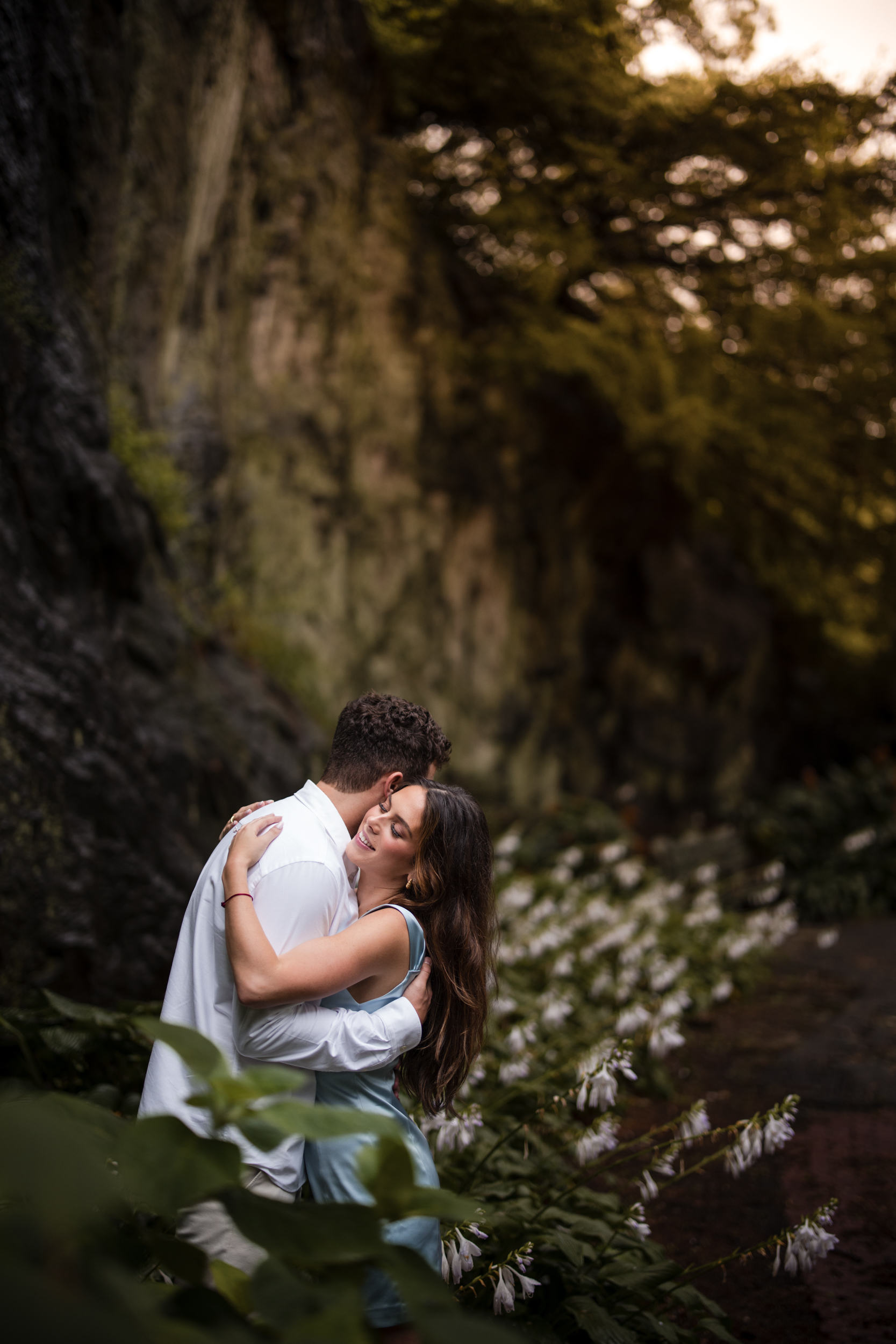 A couple stands embracing on a path lined with white flowers, surrounded by greenery and rocky cliffs, capturing the romantic essence of Fort Tryon Park engagement photos.