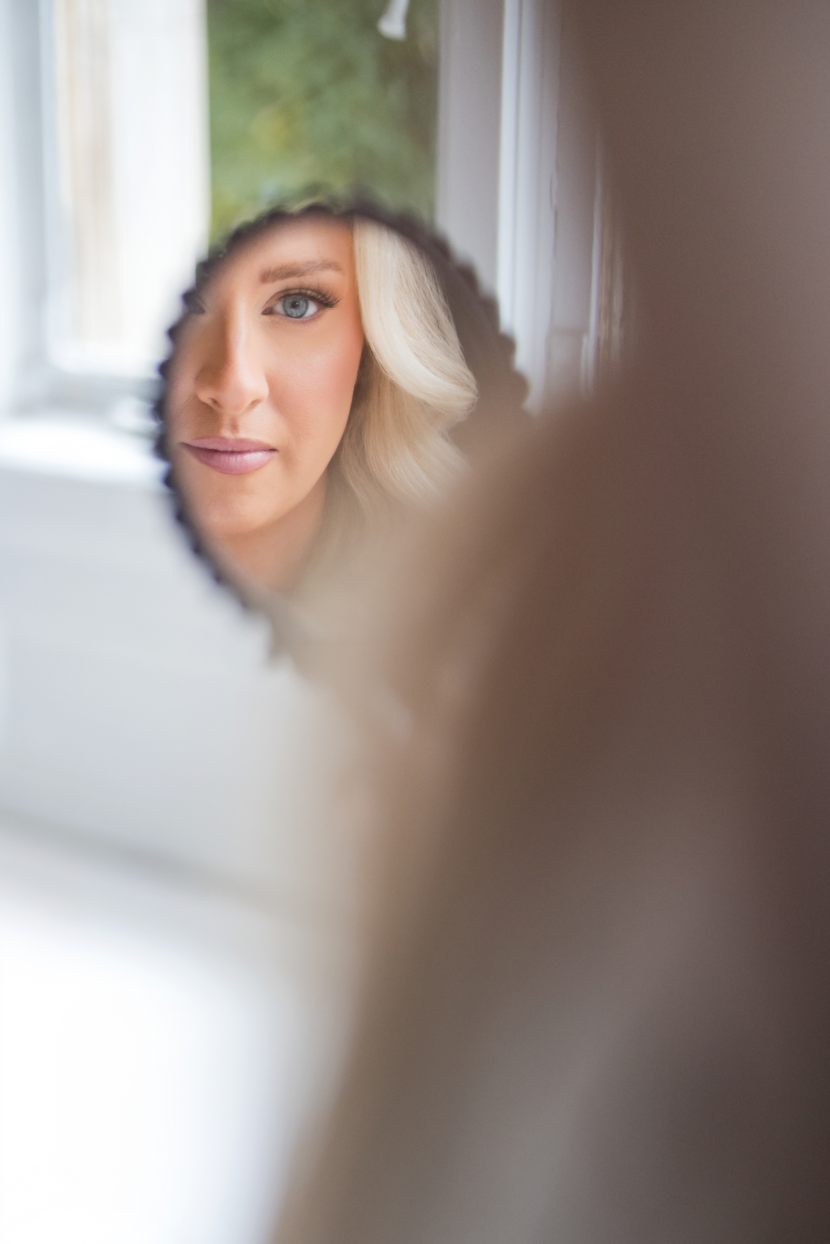 Woman’s face reflected in a small round mirror, with her features partially obscured; background is softly blurred, evoking the quiet anticipation before a Hempstead House at Sands Point Preserve wedding.