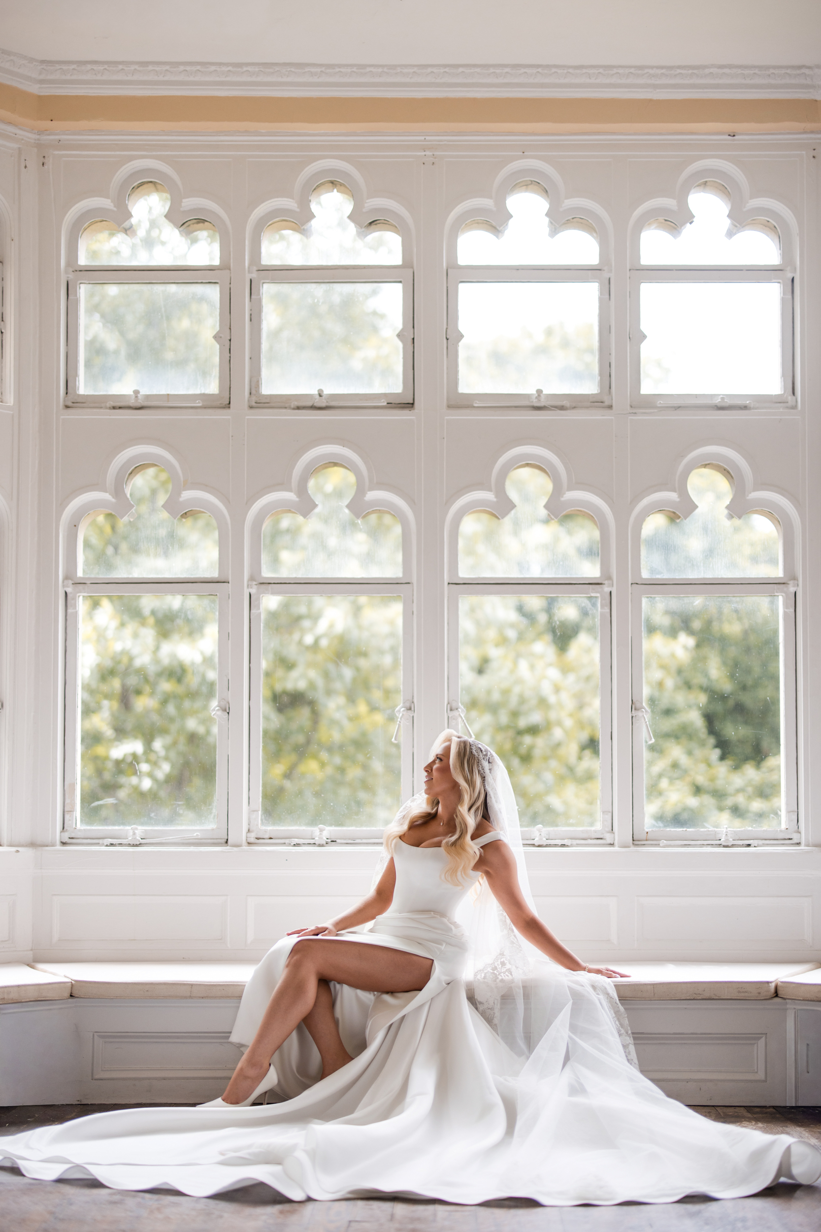 A bride in a white dress and veil sits on a windowsill in front of large arched windows at Hempstead House at Sands Point Preserve, looking to the side with one leg crossed and her train elegantly spread out.