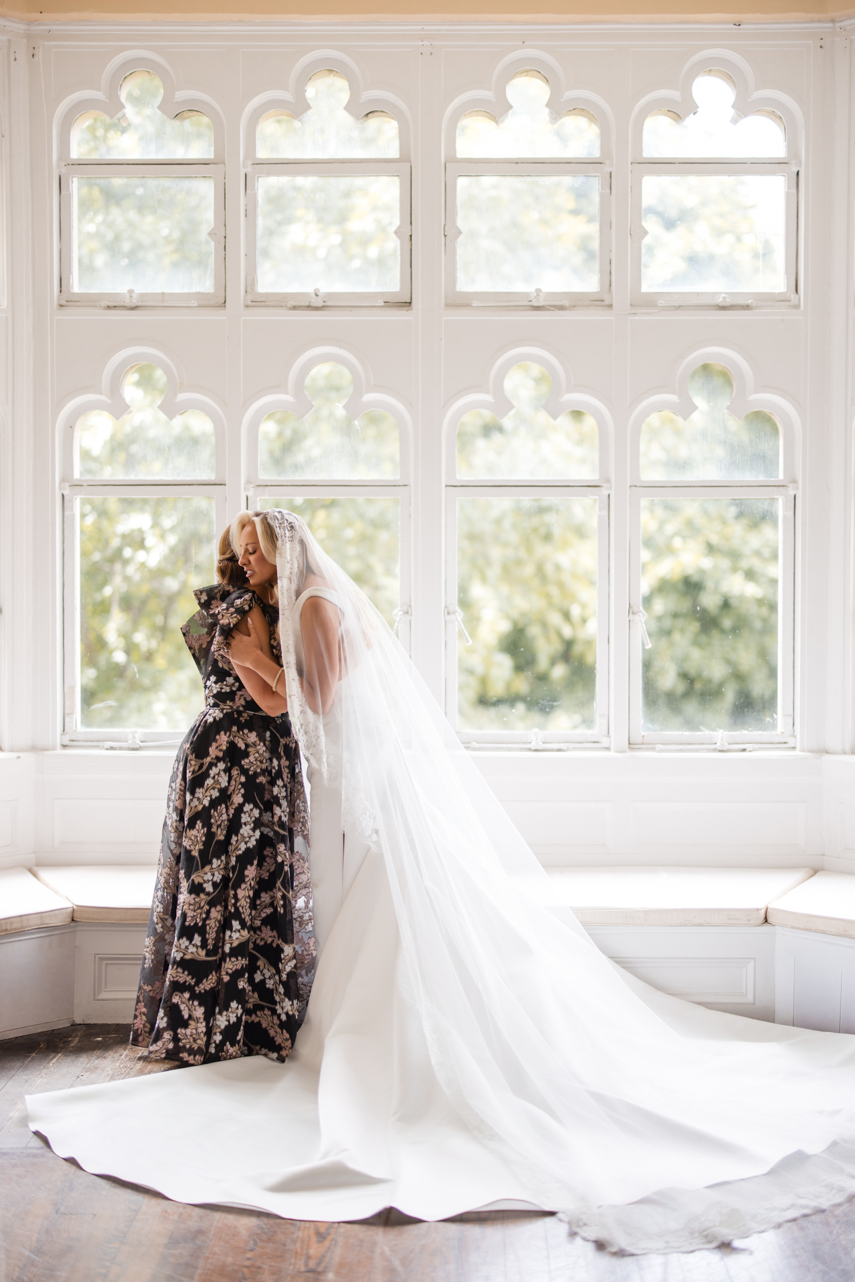 A bride in a long white dress and veil hugs a woman in a floral dress in front of tall decorative windows with light streaming in during a beautiful Hempstead House at Sands Point Preserve wedding.