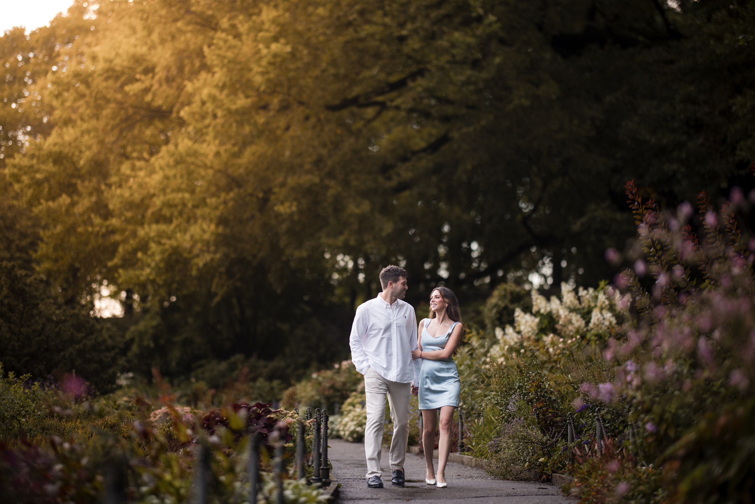 A man and a woman walk together on a garden path surrounded by greenery and flowers in Fort Tryon Park, with sunlight filtering through the trees—capturing the essence of romantic engagement photos.