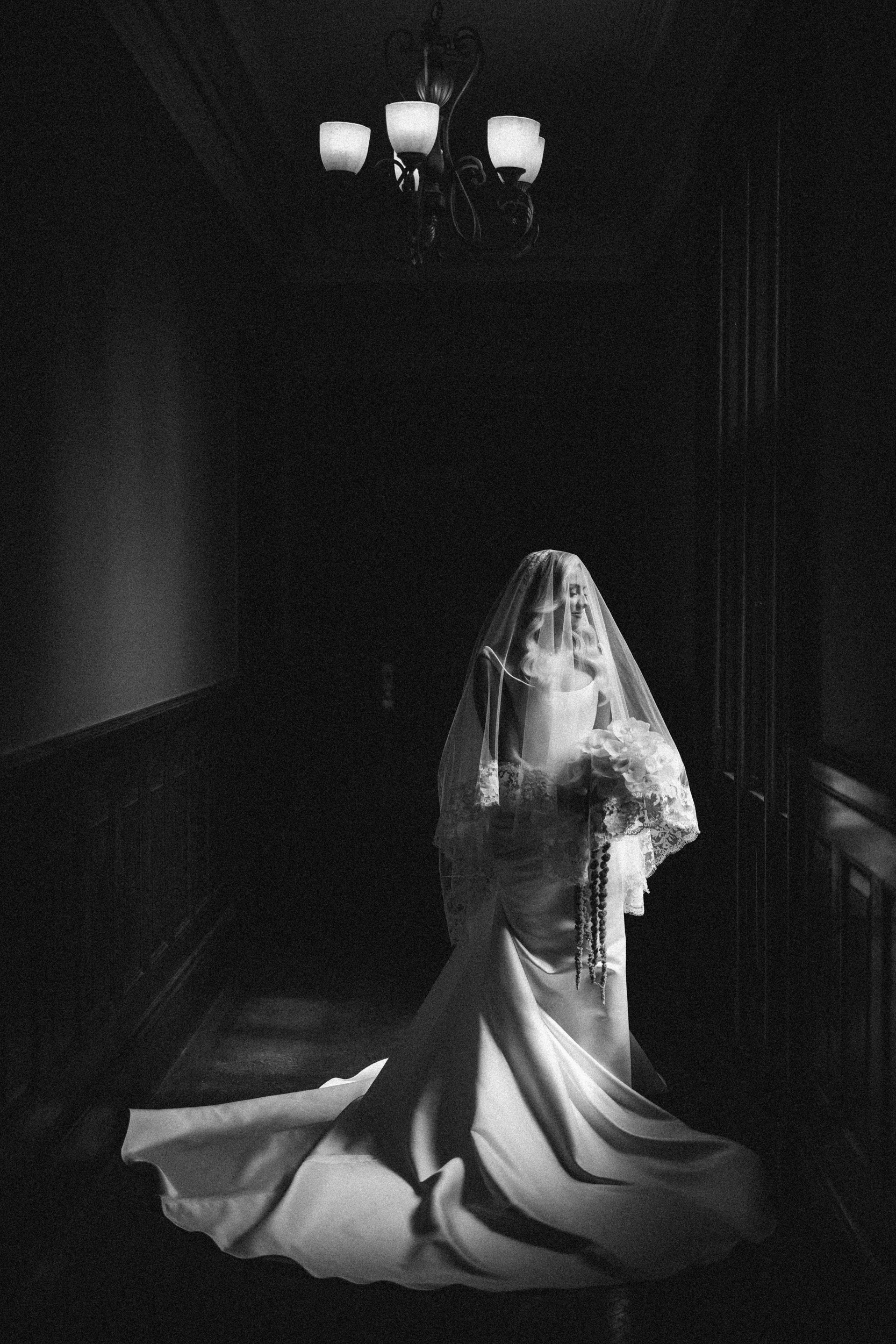 A bride in a long gown and veil stands alone in a dimly lit hallway of Hempstead House at Sands Point Preserve, holding a bouquet. Black and white photo.