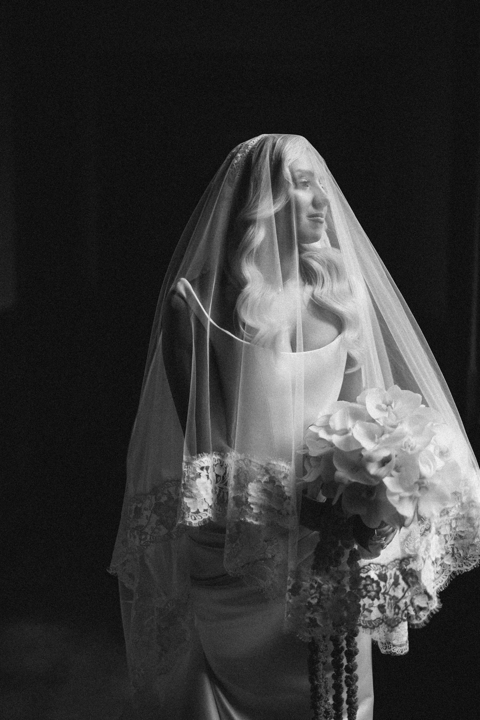 A bride in a lace-trimmed veil and gown holds a bouquet of flowers, looking to the side in a softly lit, black-and-white photo captured during a romantic Hempstead House at Sands Point Preserve wedding.