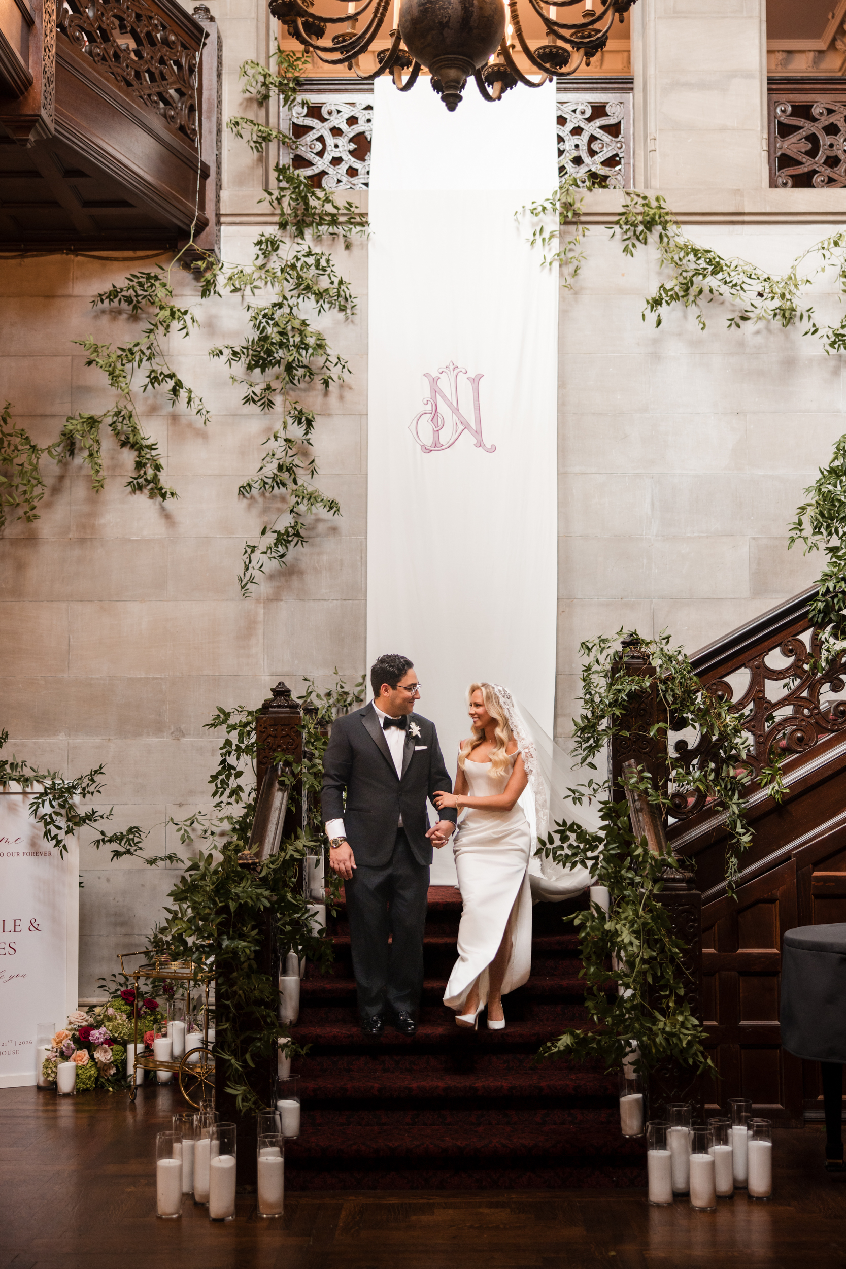 A bride and groom walk down a staircase decorated with greenery and candles in an elegant indoor setting, capturing the timeless romance of a Hempstead House at Sands Point Preserve wedding.