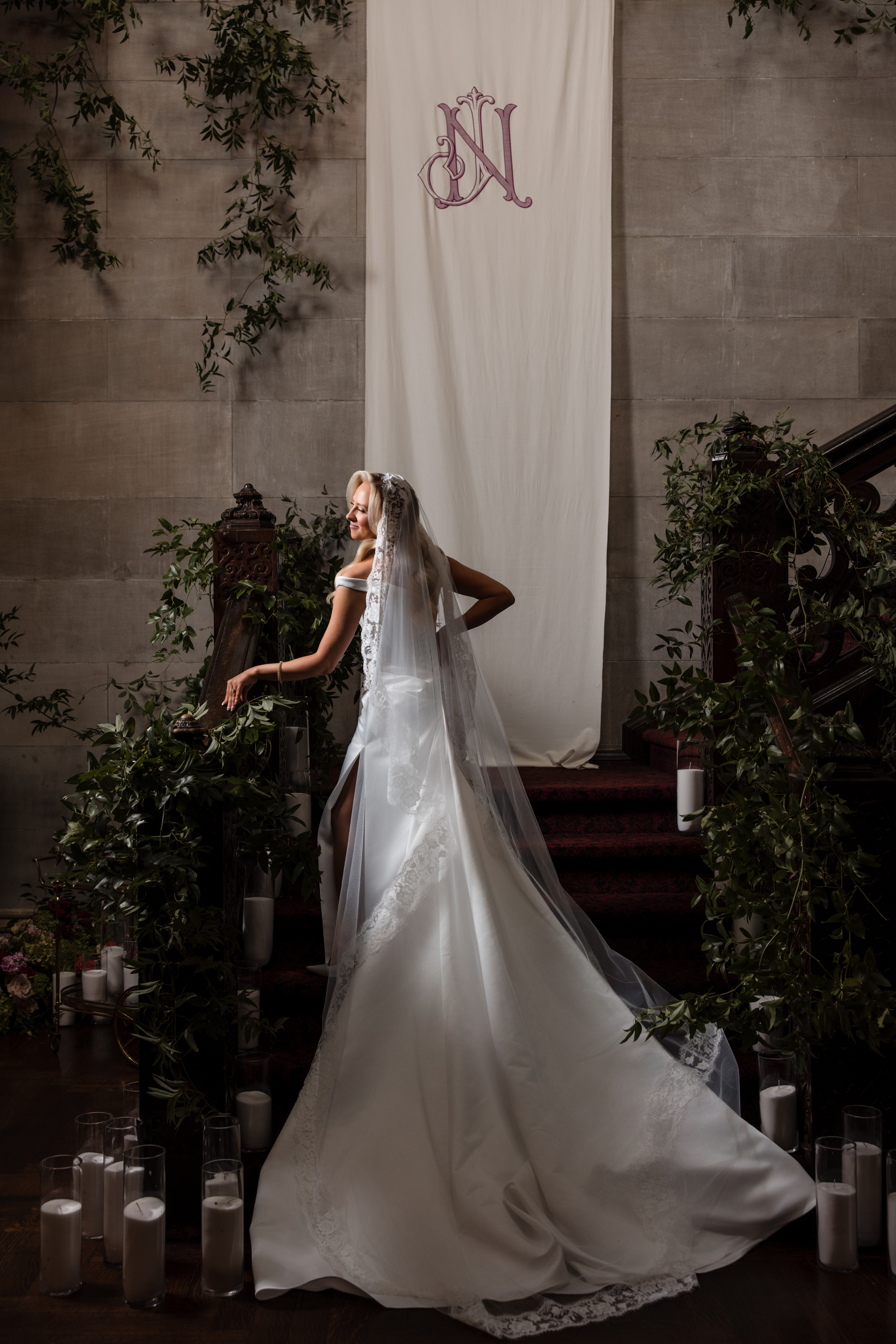 A bride in a white gown and veil stands on a decorated staircase with greenery and candles, in front of a banner with a monogram, capturing the elegance of a Hempstead House at Sands Point Preserve wedding.