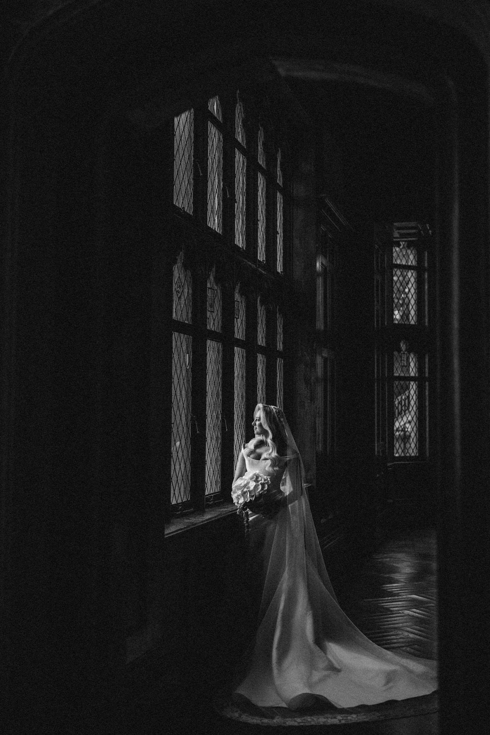 A bride in a long gown and veil stands by a tall window in a dimly lit, ornate hallway at Hempstead House at Sands Point Preserve, holding a bouquet of flowers.