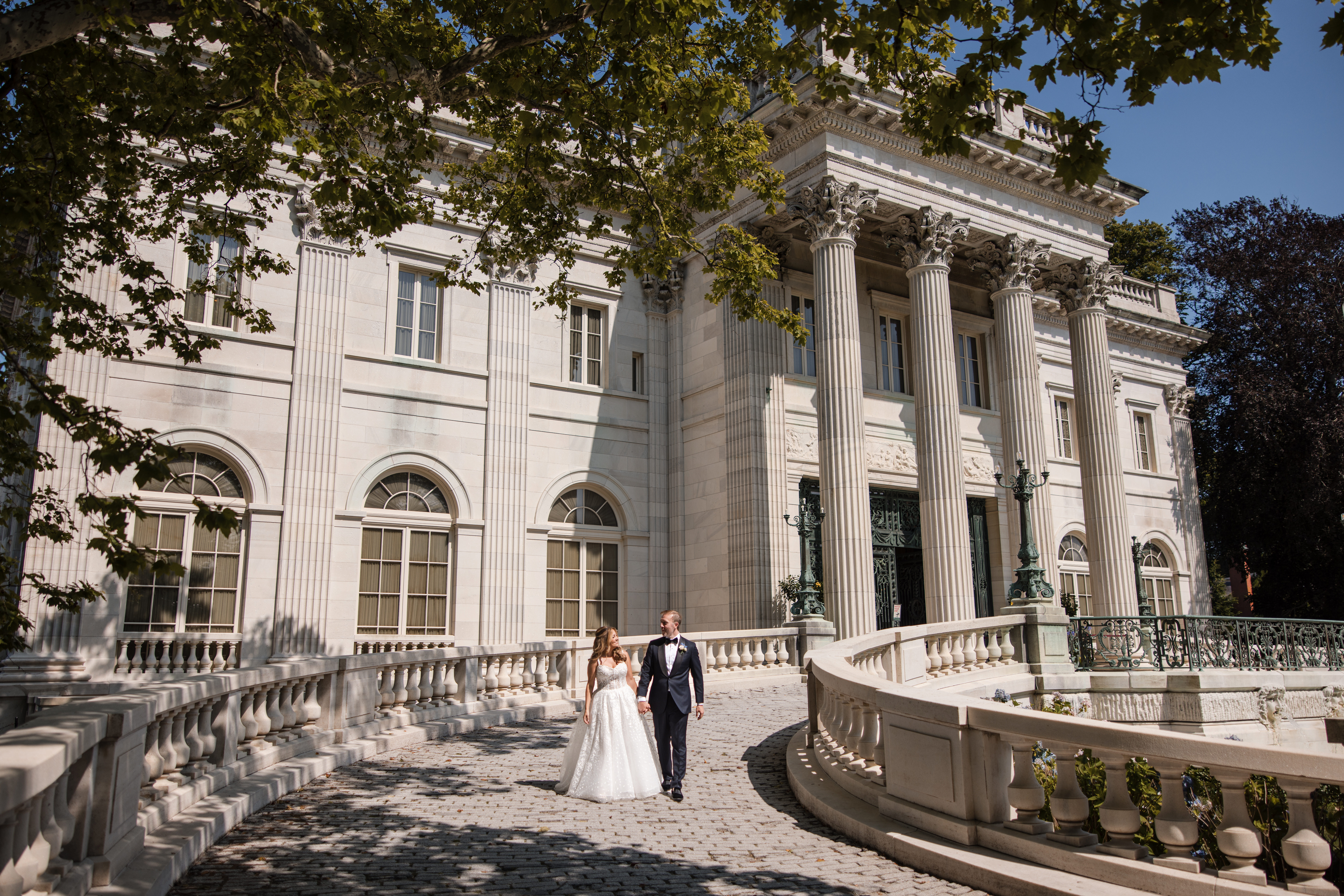A bride and groom walk together on a curved stone path outside a large, ornate building with tall columns and arched windows on a sunny day, capturing timeless Marble House portraits reminiscent of an elegant Regatta Place wedding.