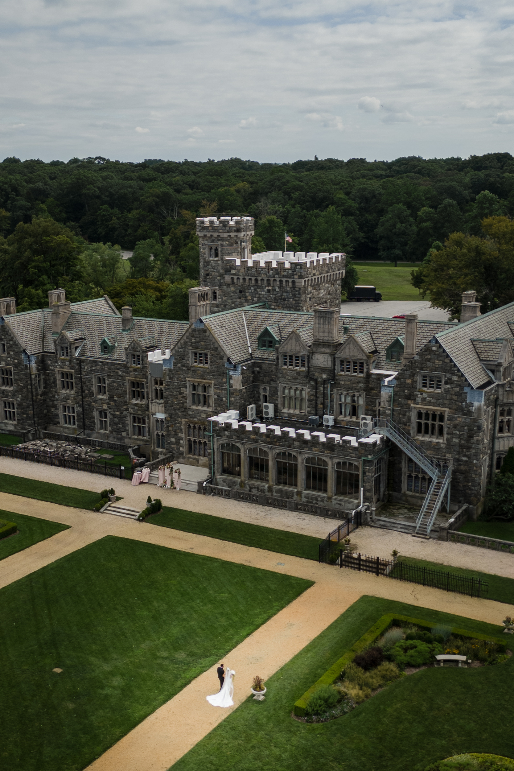 A bride and groom stand together on a walkway in front of a large stone castle surrounded by manicured gardens and greenery, capturing the elegance of a Hempstead House at Sands Point Preserve wedding.