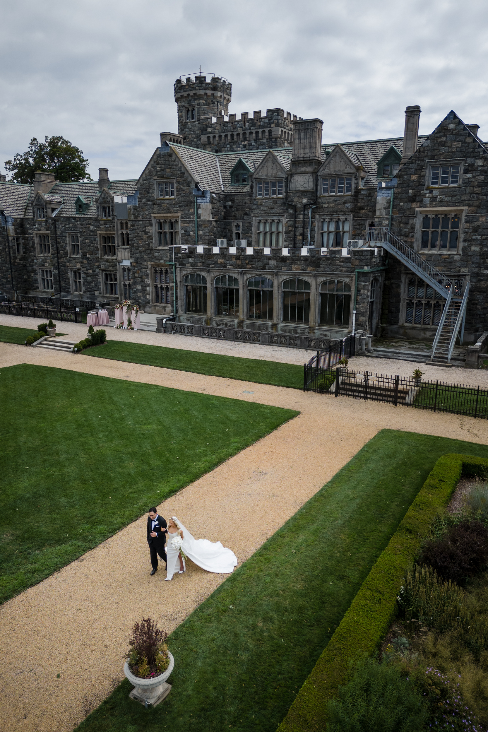 A bride and groom walk hand in hand along a gravel path outside the grand Hempstead House at Sands Point Preserve, its castle-like architecture framed by manicured lawns and cloudy skies above—a picturesque setting for their wedding day.