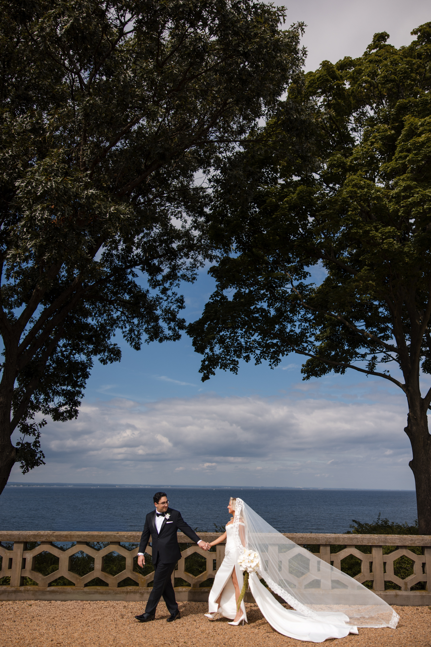 A bride and groom walk hand in hand outdoors between two large trees, with ocean and cloudy sky in the background. The bride’s veil flows behind her, capturing the romantic atmosphere of a Hempstead House at Sands Point Preserve wedding.