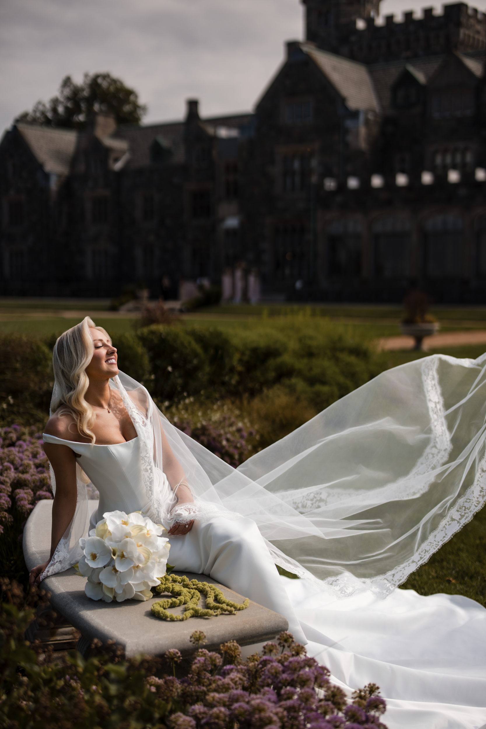 A bride in a white gown and veil sits outdoors on a stone bench, holding a bouquet of white flowers, with the grand Hempstead House at Sands Point Preserve wedding venue standing elegantly in the background.
