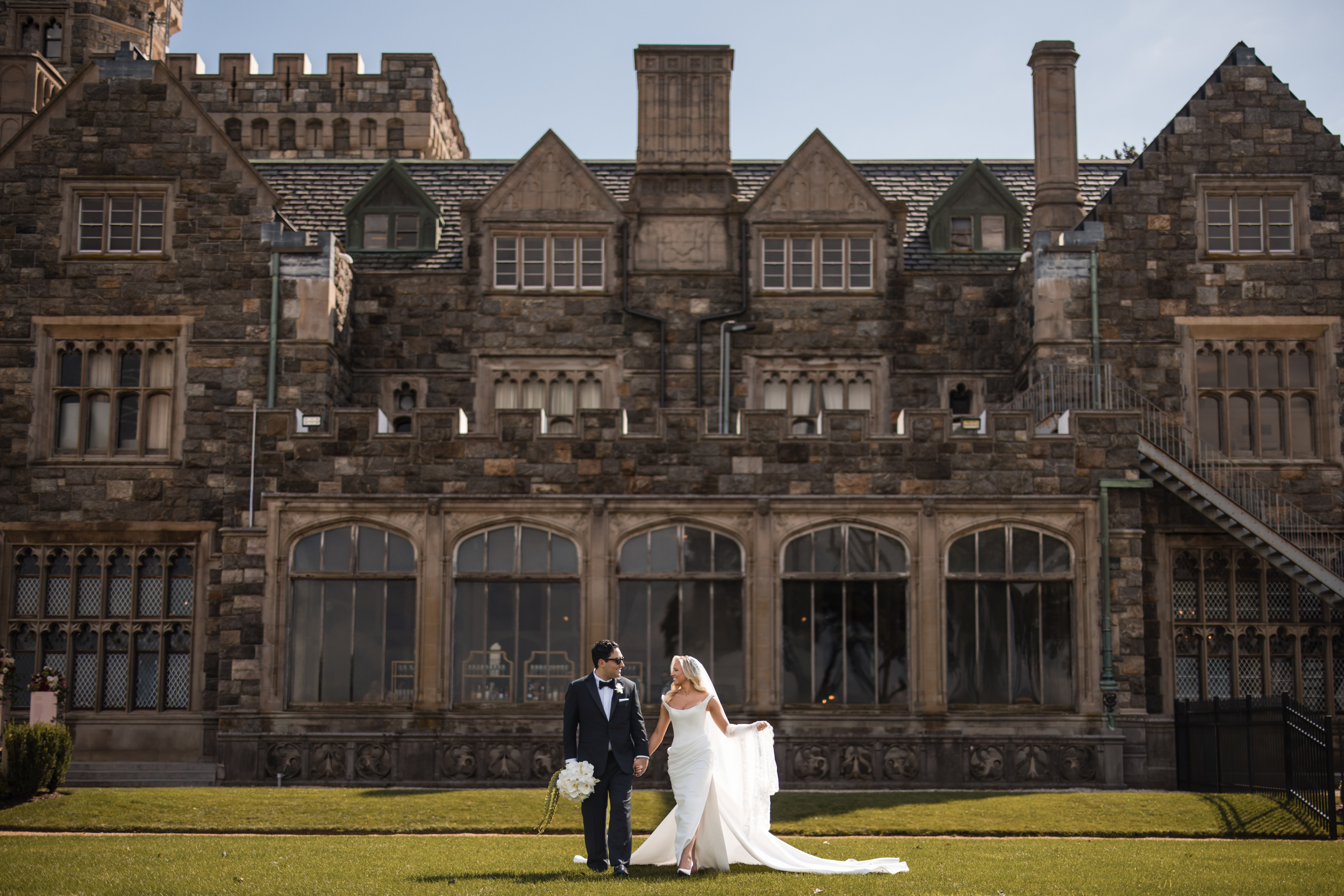 A bride and groom in formal wedding attire stand holding hands on a lawn in front of the grand Hempstead House at Sands Point Preserve, its large stone walls, tall windows, and chimneys providing a stunning backdrop to their wedding day. A bride and groom in formal wedding attire stand holding hands on a lawn in front of the grand Hempstead House at Sands Point Preserve, its large stone walls, tall windows, and chimneys providing a stunning backdrop to their wedding day.