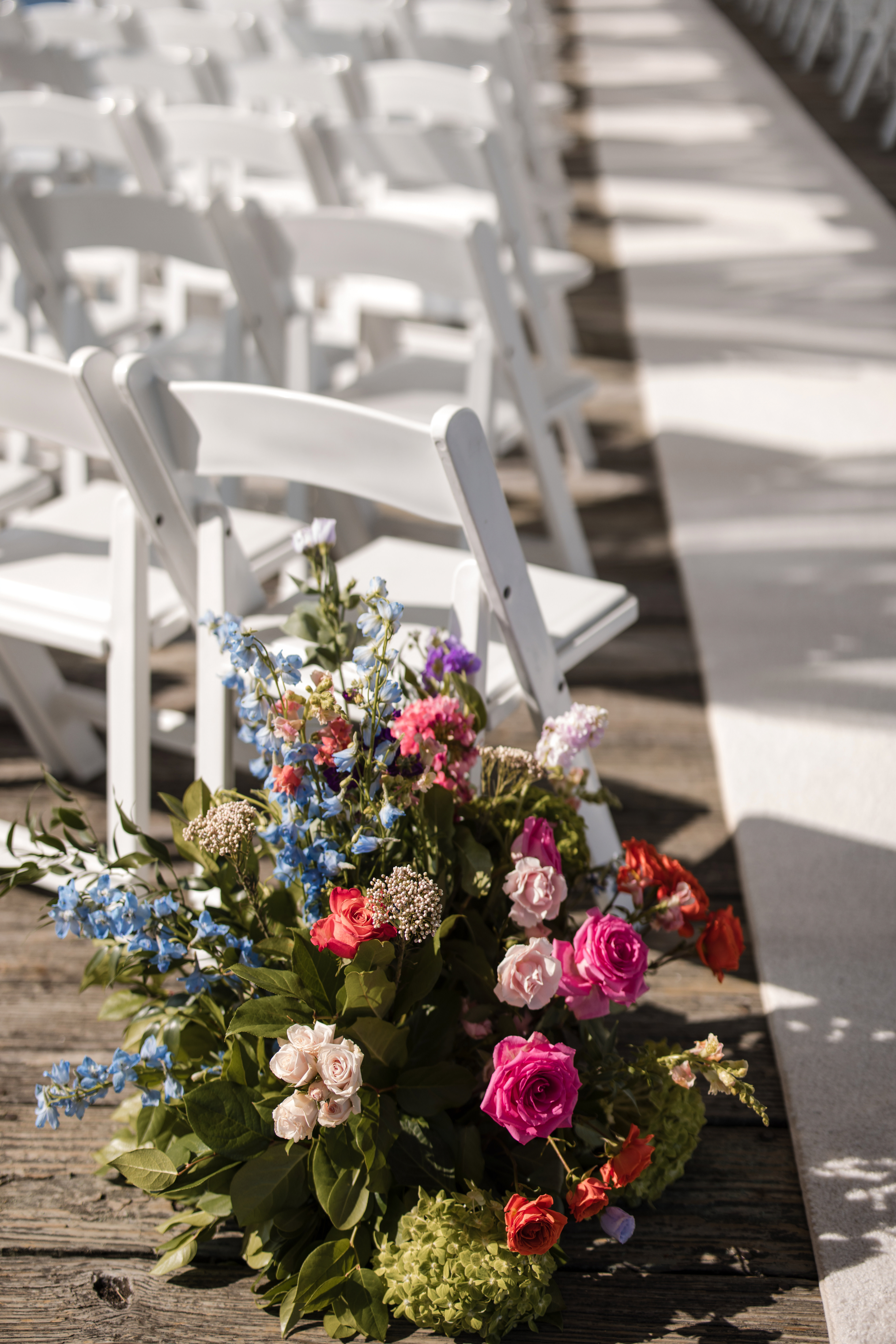 A colorful floral arrangement sits beside white folding chairs arranged in rows along a wooden aisle at an outdoor Regatta Place wedding.
