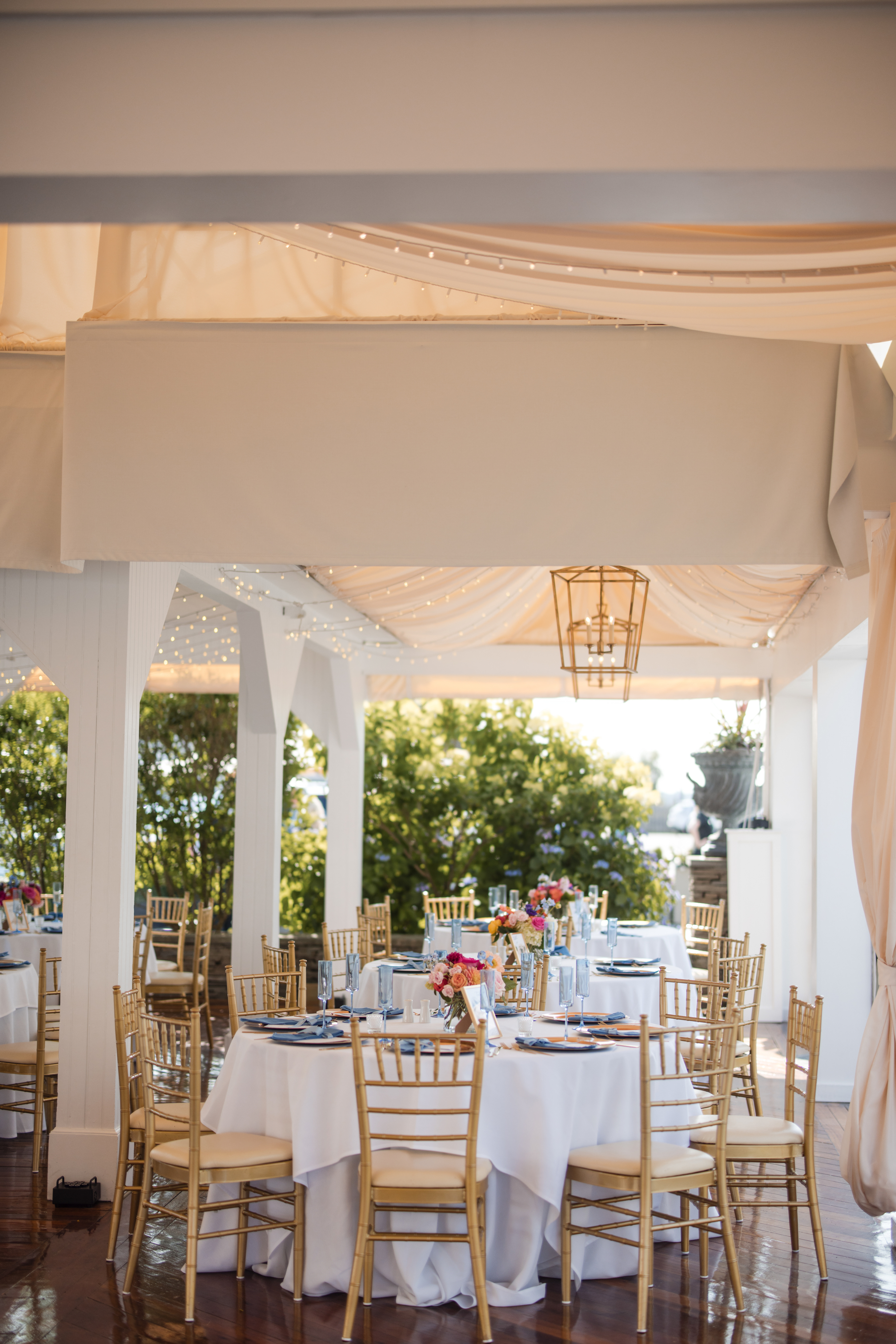 Elegant outdoor dining area with round tables, gold chairs, white tablecloths, and floral centerpieces under a draped canopy with string lights—perfect for a memorable Regatta Place wedding.