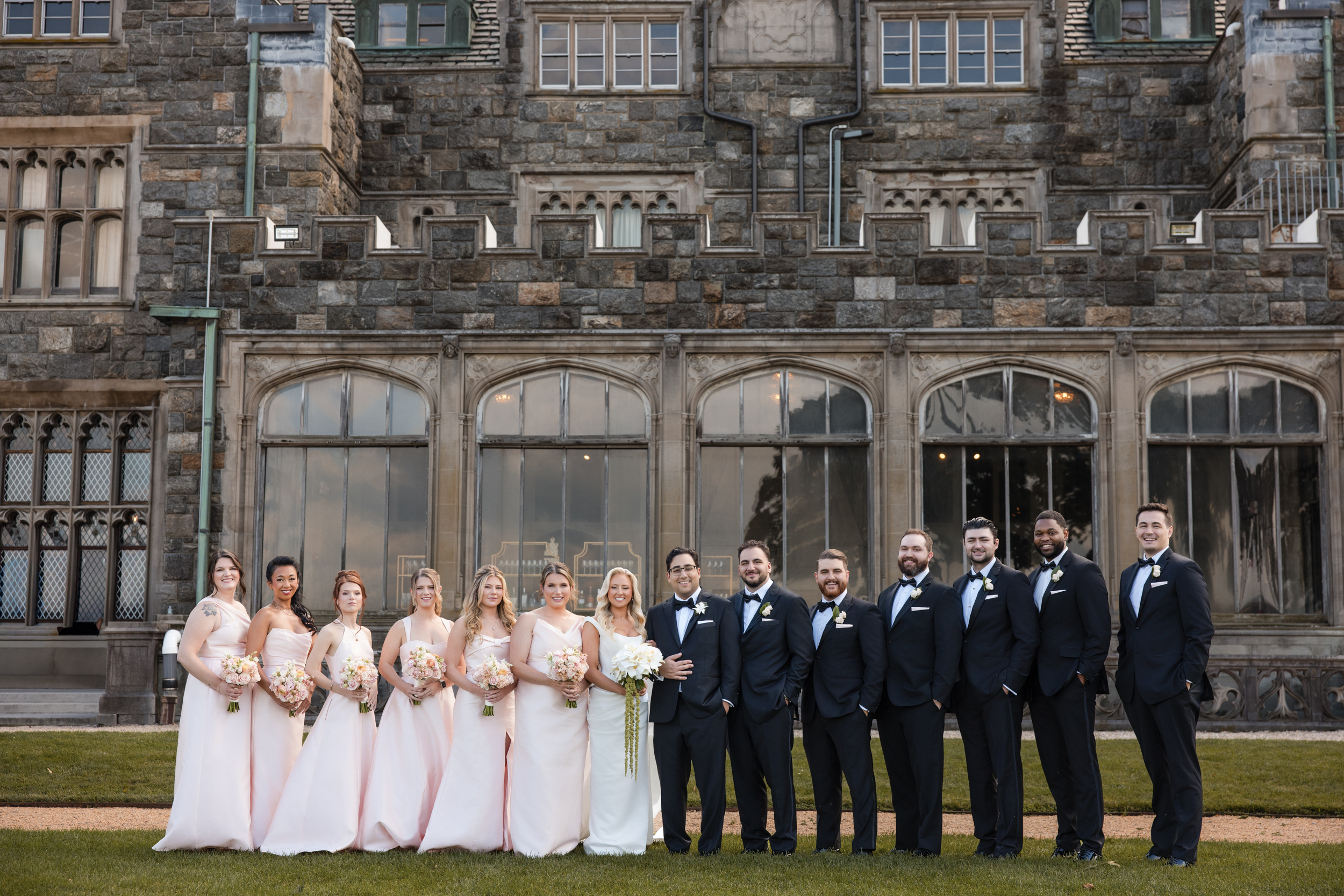 A bride, groom, and wedding party pose in formal attire outside Hempstead House at Sands Point Preserve, a large stone building with tall windows and a manicured lawn in front. A bride, groom, and wedding party pose in formal attire outside Hempstead House at Sands Point Preserve, a large stone building with tall windows and a manicured lawn in front.