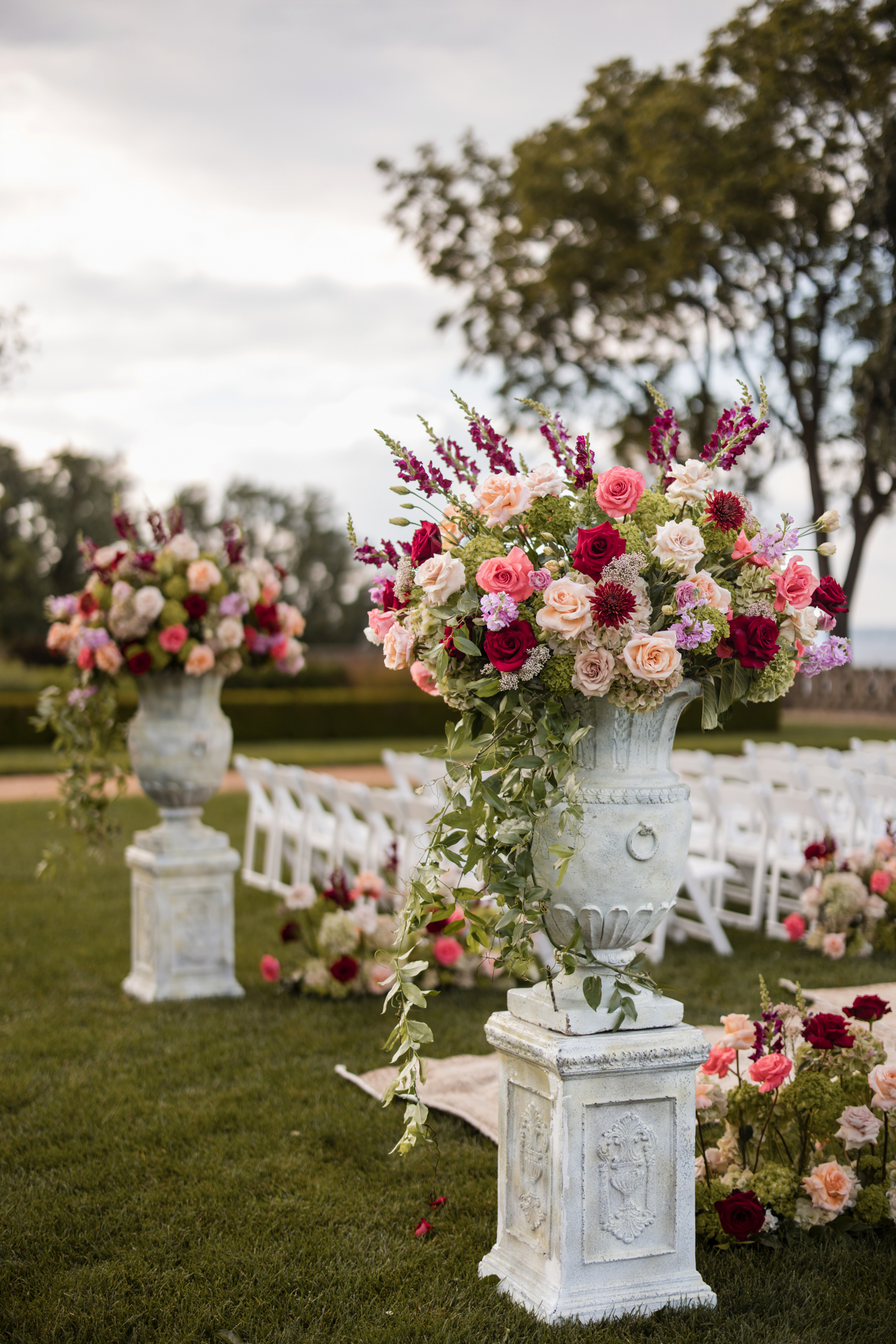 Large floral arrangements in white stone urns line an outdoor aisle with white chairs and scattered flowers, set on a grassy lawn with trees in the background—capturing the timeless elegance of a Hempstead House at Sands Point Preserve wedding.