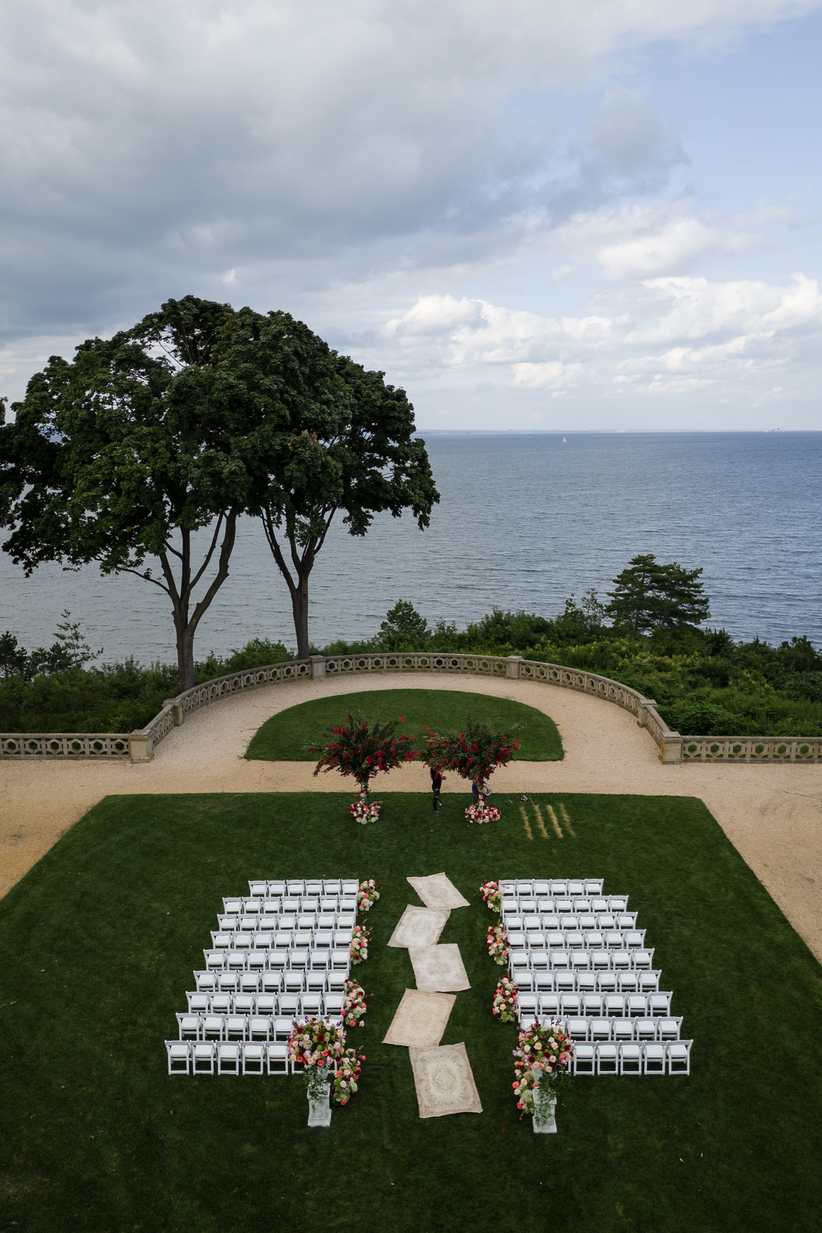Rows of white chairs arranged for an outdoor Hempstead House at Sands Point Preserve wedding ceremony on a lawn, with flower decorations and a view of the ocean and trees in the background under a cloudy sky.