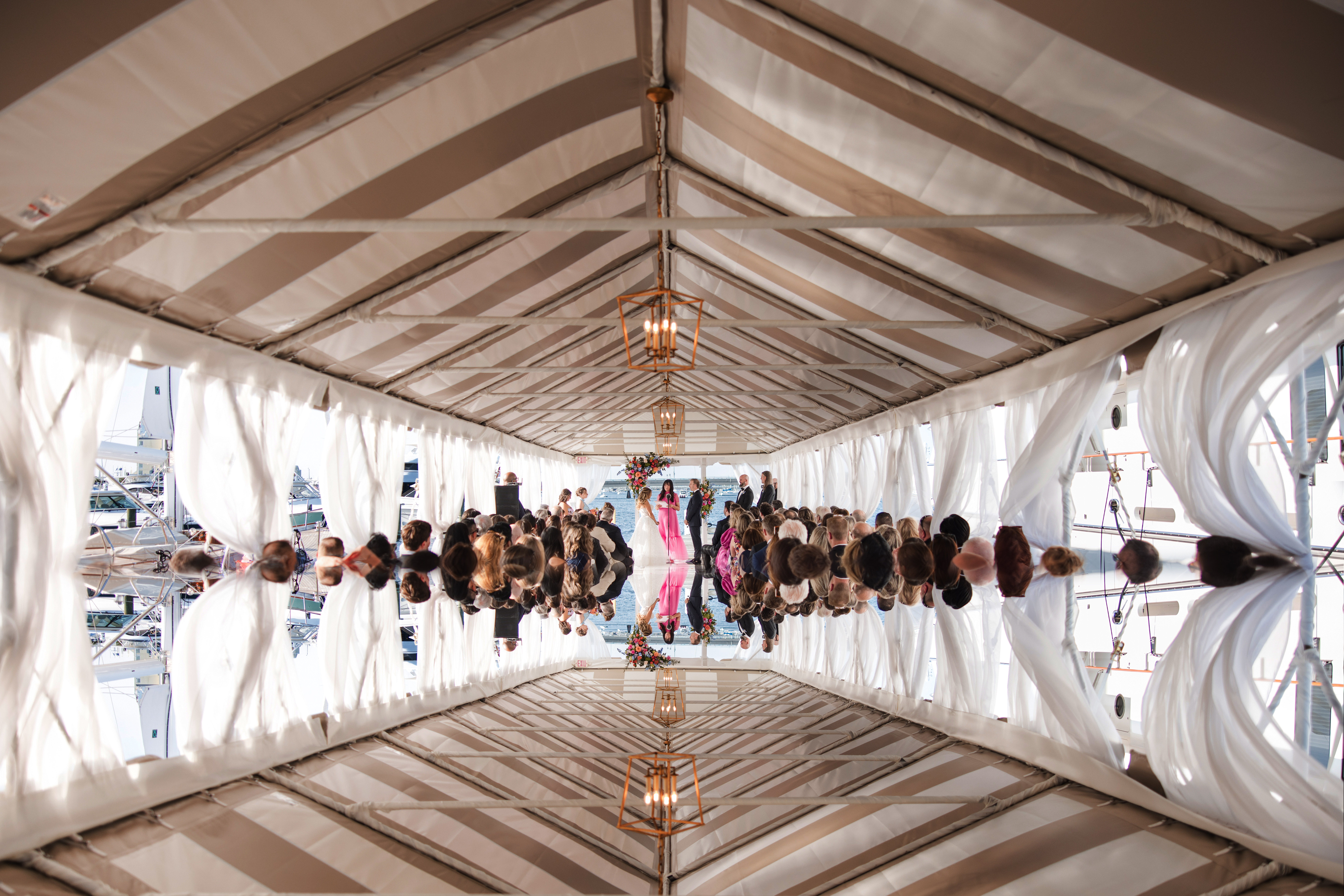 A Regatta Place wedding ceremony takes place under a white tent with guests seated on both sides, draped curtains, and a chandelier hanging overhead. The image is mirrored vertically, creating a symmetrical effect.