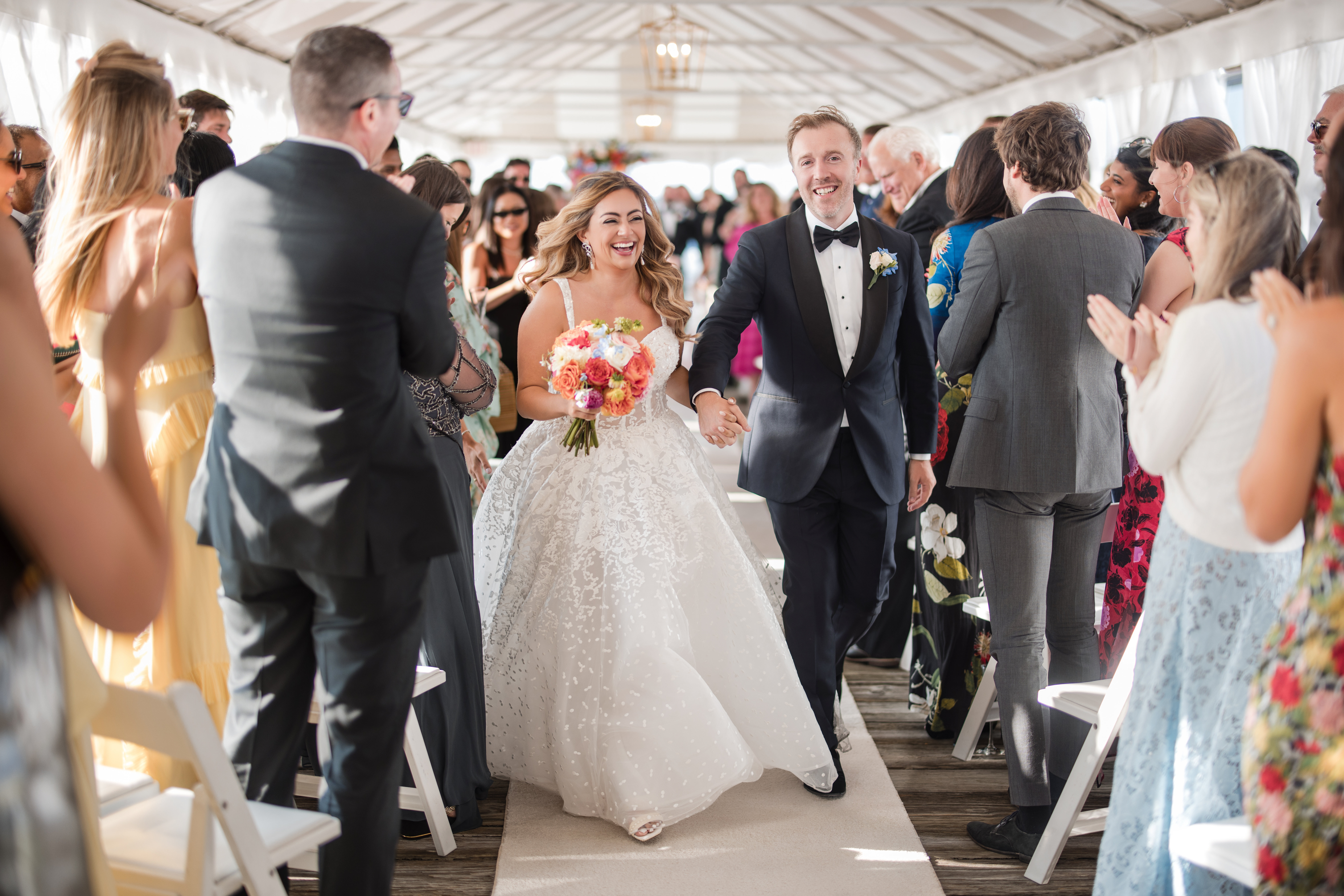 A bride and groom walk down the aisle together at their Regatta Place wedding, smiling and holding hands, while wedding guests stand and applaud on both sides.
