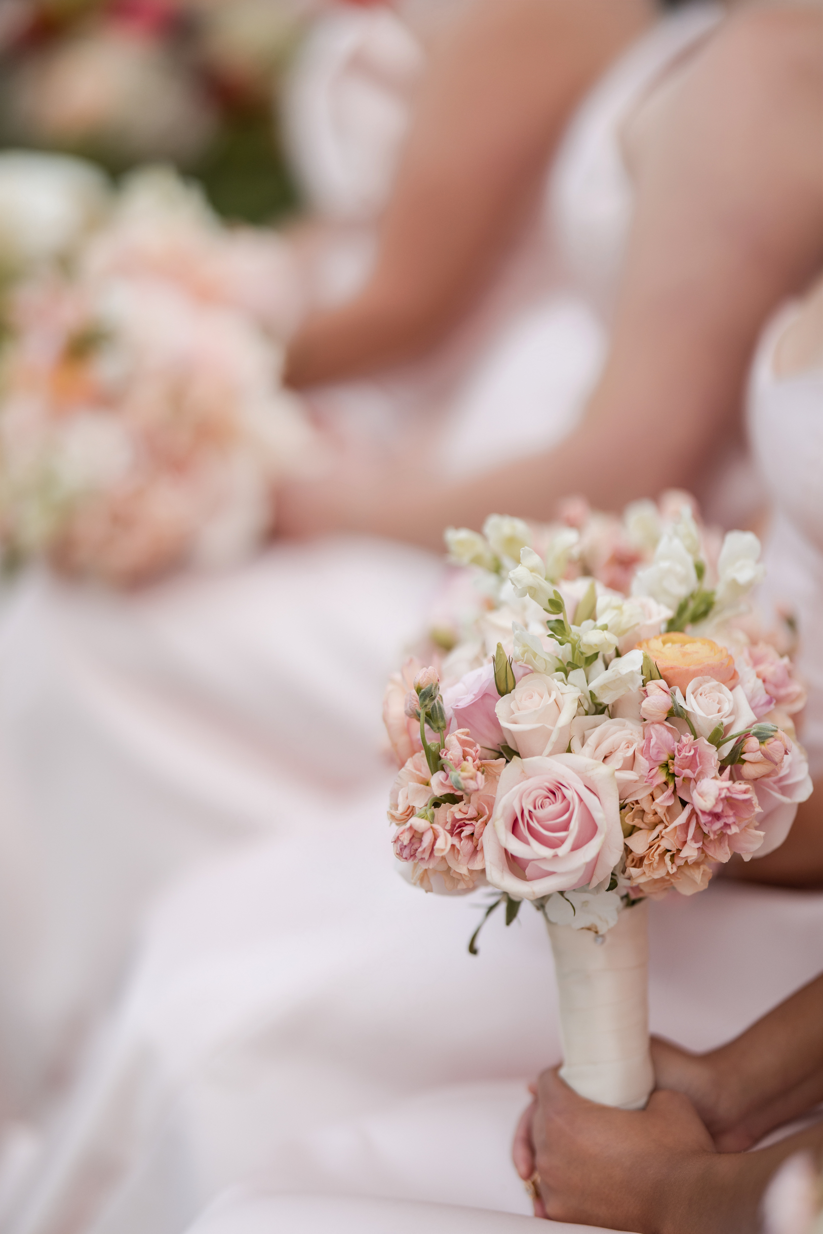 Close-up of hands holding a bouquet of pink and white flowers, with others in the background, all dressed in light-colored formal attire at a beautiful Hempstead House at Sands Point Preserve wedding.