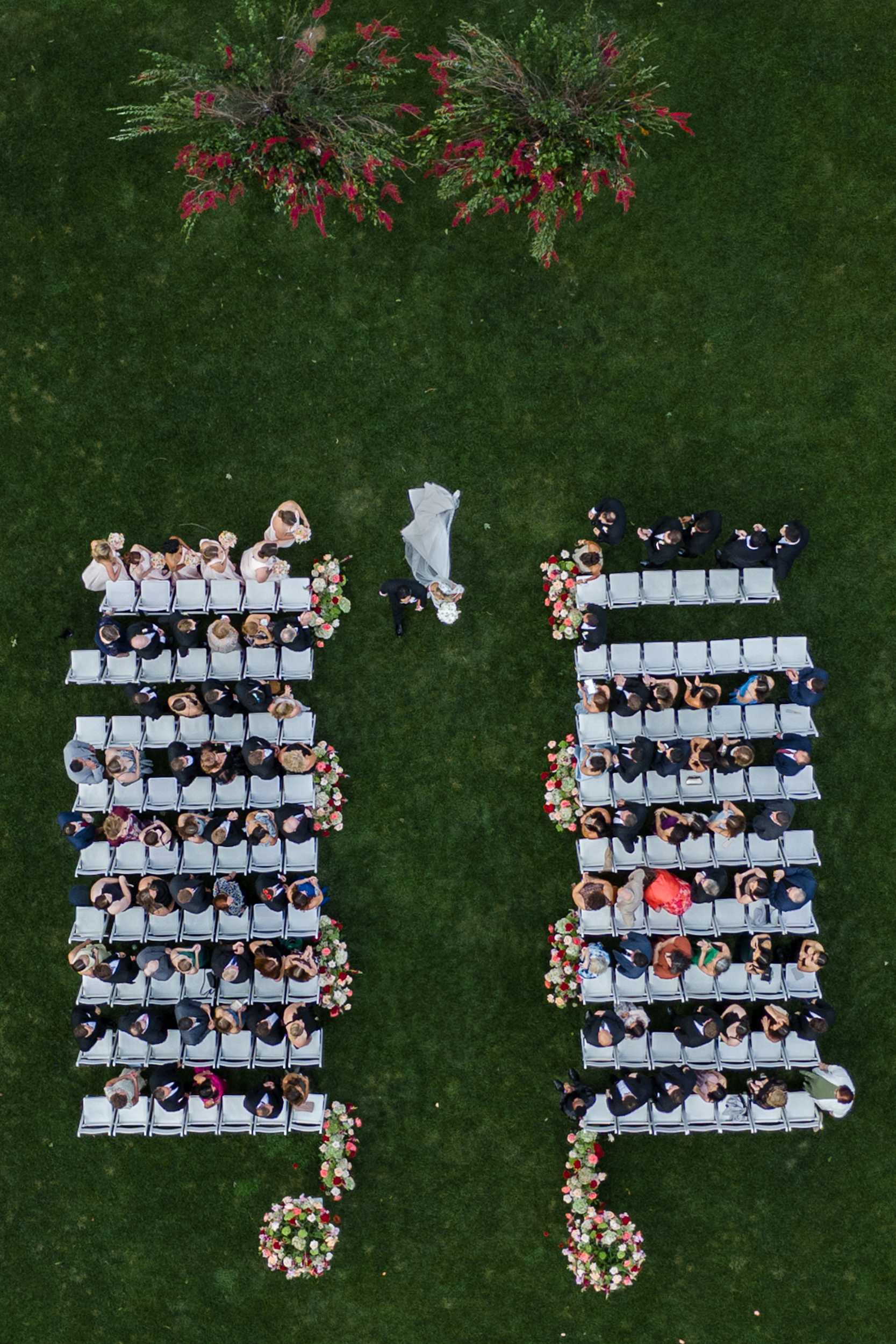 Aerial view of an outdoor Hempstead House at Sands Point Preserve wedding ceremony with guests seated in two groups, a couple walking down the aisle, and floral decorations on green grass.