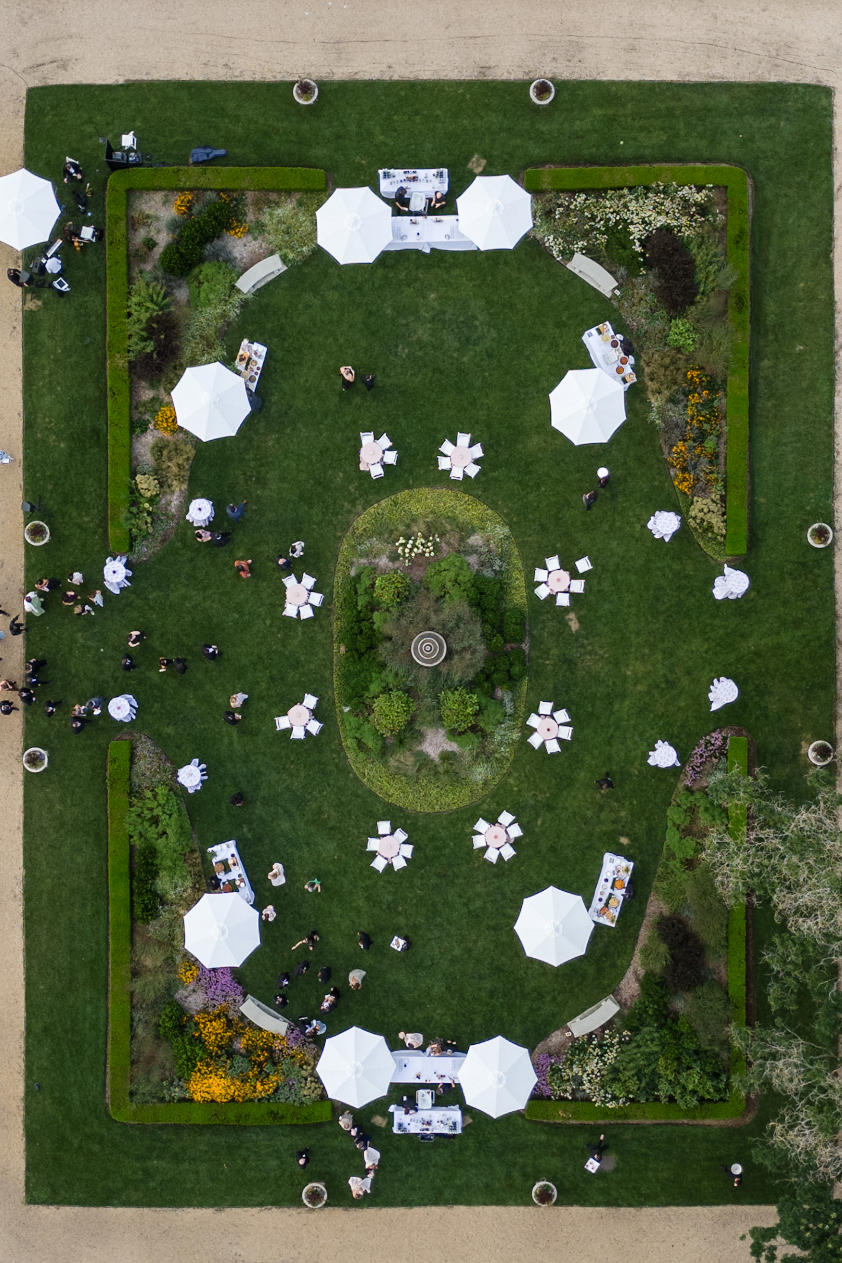 Aerial view of a rectangular garden with white umbrellas, tables, and people gathered on the grass, surrounded by flowerbeds and pathways—perfectly capturing the elegant atmosphere of a Hempstead House at Sands Point Preserve wedding.