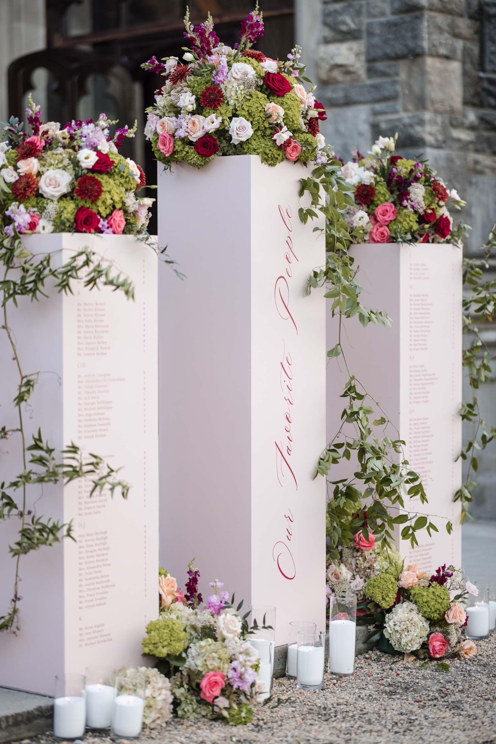 Three tall pink pedestals with guest names in lists, decorated with floral arrangements and greenery, surrounded by candles and flowers, stand in front of a stone building—creating a romantic welcome for a Hempstead House at Sands Point Preserve wedding.