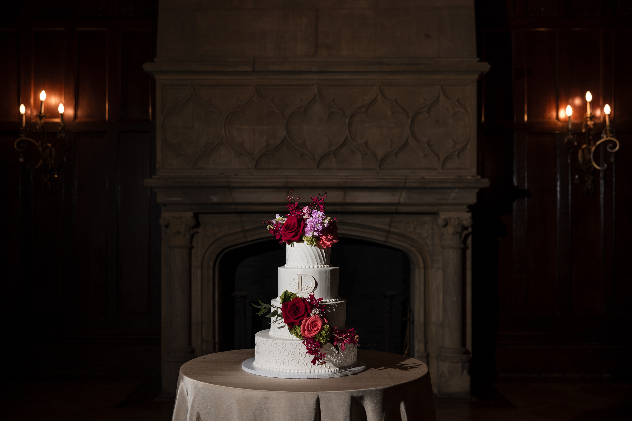 Three-tiered white cake with red and purple flowers on a round table in front of a large stone fireplace, illuminated by side wall sconces—the perfect centerpiece for a romantic Hempstead House at Sands Point Preserve wedding. Three-tiered white cake with red and purple flowers on a round table in front of a large stone fireplace, illuminated by side wall sconces—the perfect centerpiece for a romantic Hempstead House at Sands Point Preserve wedding.