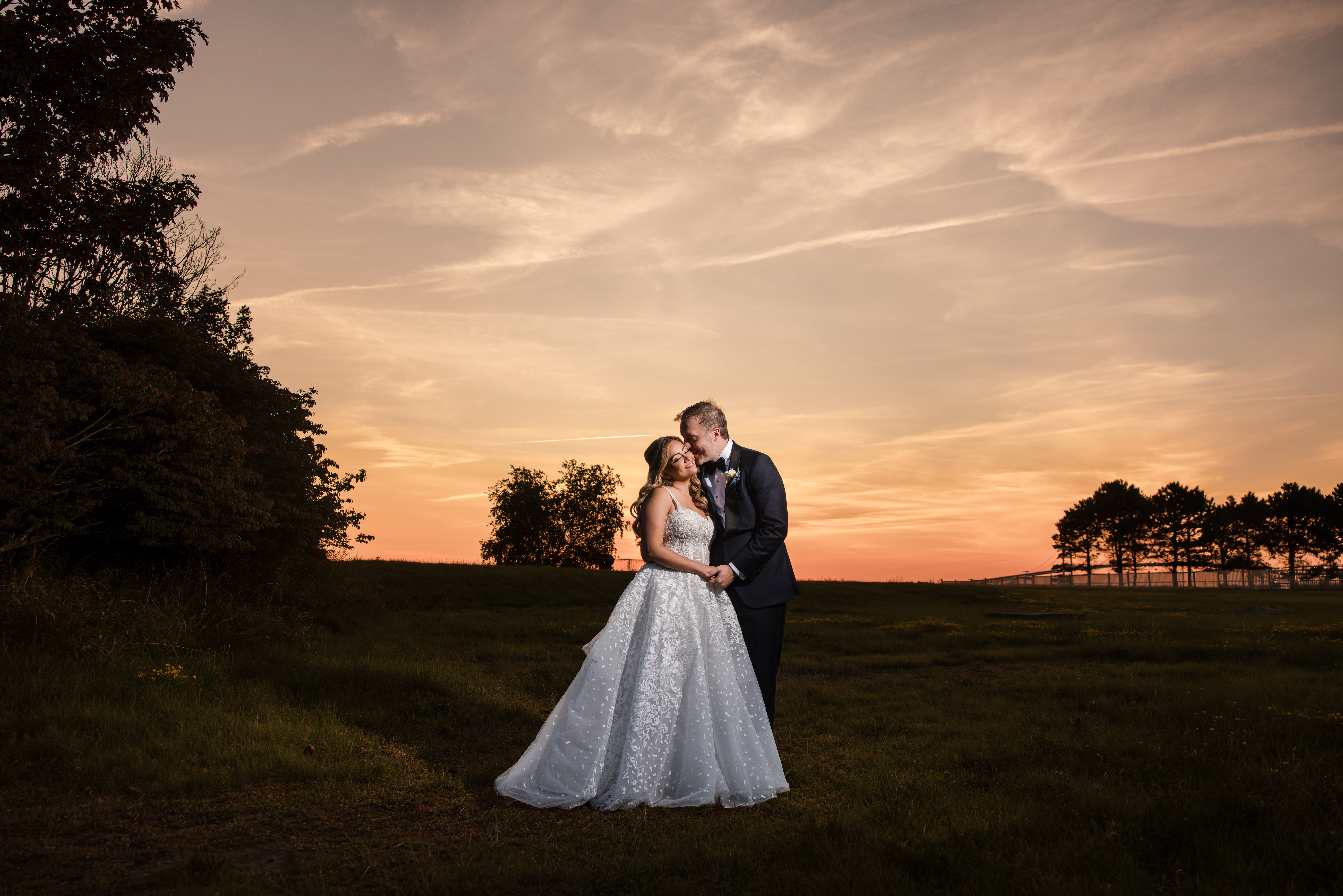 A bride and groom stand together in a field at sunset, holding hands and embracing, with trees and a colorful sky in the background, capturing the romantic ambiance of a Regatta Place wedding.