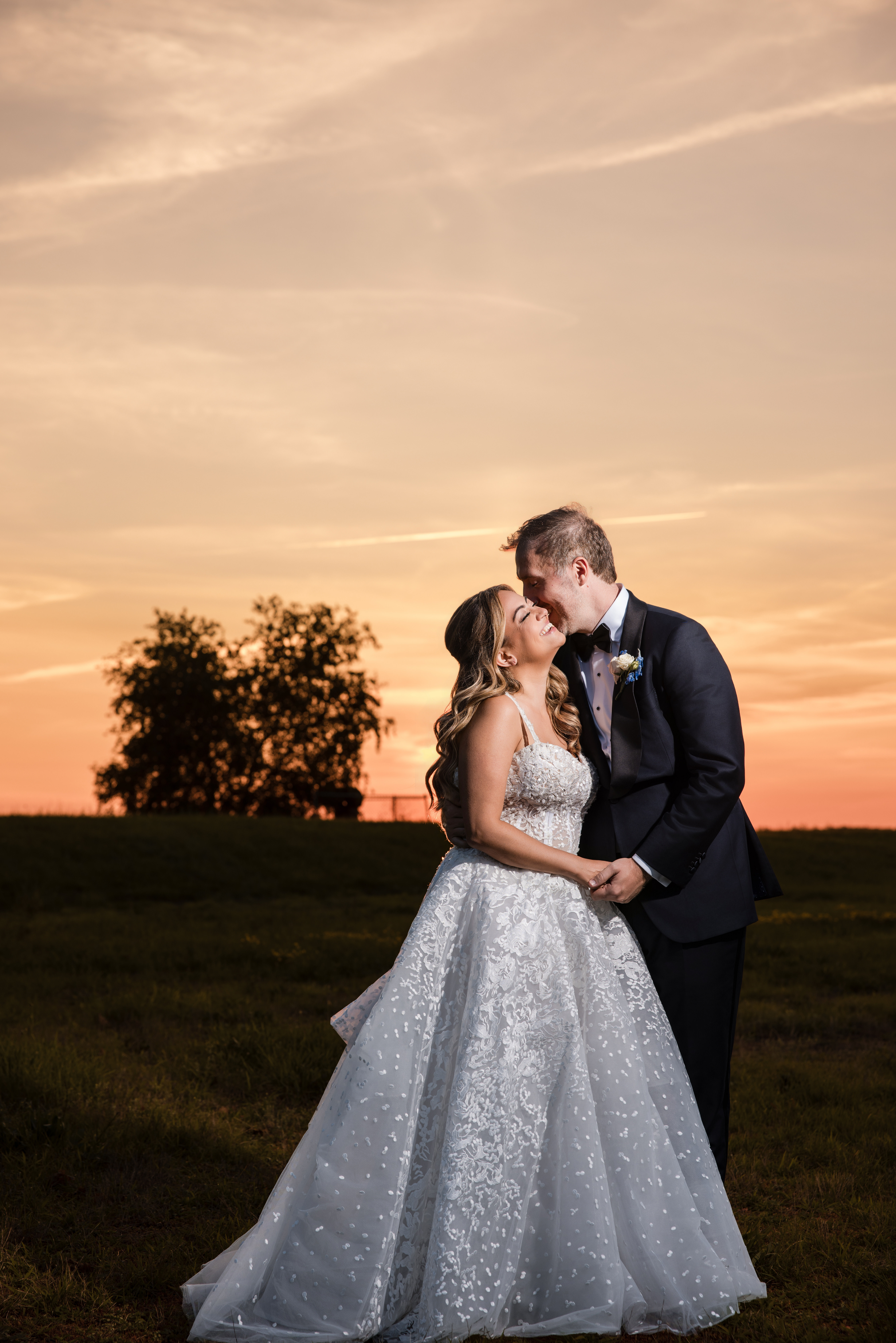 A bride and groom in formal wedding attire stand outdoors at sunset, holding hands and sharing a kiss, with trees and an orange sky in the background, capturing the romantic ambiance of a Regatta Place wedding.