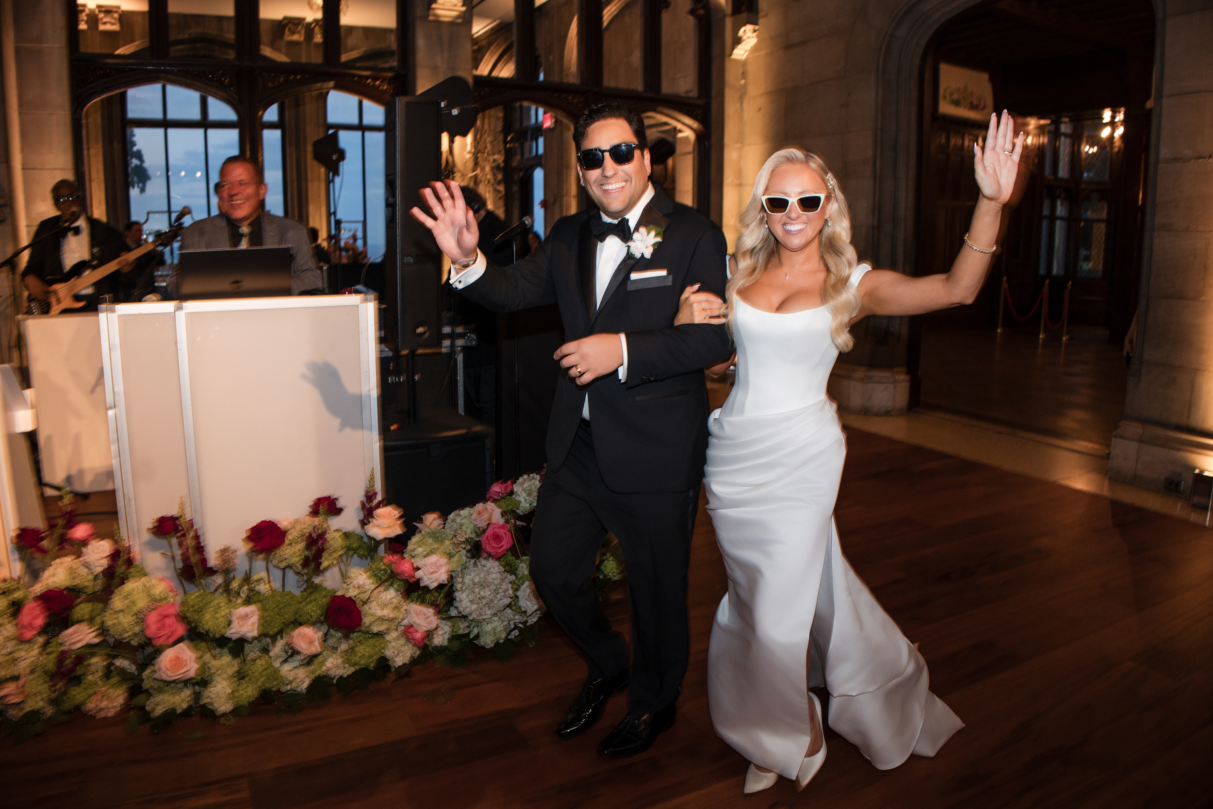 A bride and groom, both wearing sunglasses, smile and wave as they enter their elegant Hempstead House at Sands Point Preserve wedding reception, a formal indoor celebration beautifully decorated with flowers. A bride and groom, both wearing sunglasses, smile and wave as they enter their elegant Hempstead House at Sands Point Preserve wedding reception, a formal indoor celebration beautifully decorated with flowers.