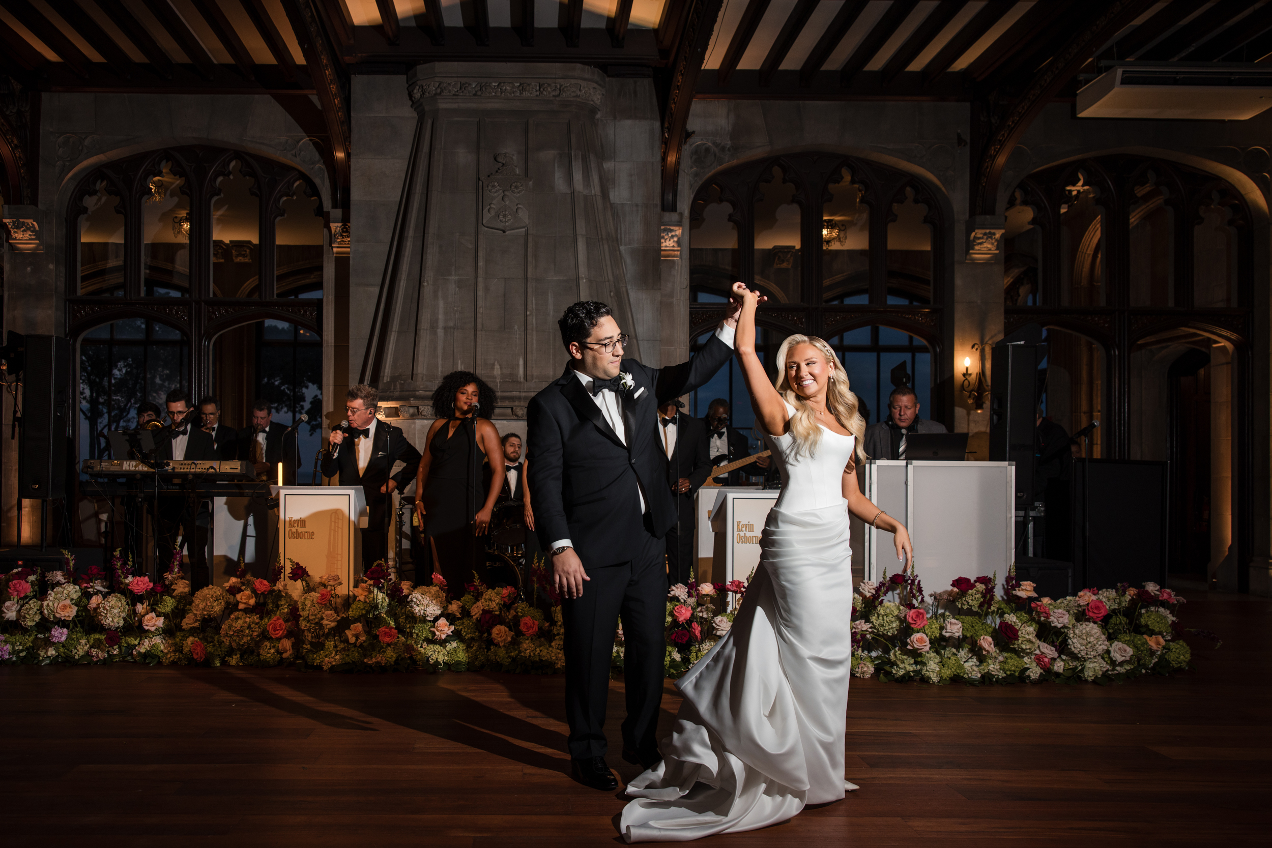 A bride and groom dance together in formal attire at their Hempstead House at Sands Point Preserve wedding reception, with a live band performing in the background and floral arrangements on the stage. A bride and groom dance together in formal attire at their Hempstead House at Sands Point Preserve wedding reception, with a live band performing in the background and floral arrangements on the stage.